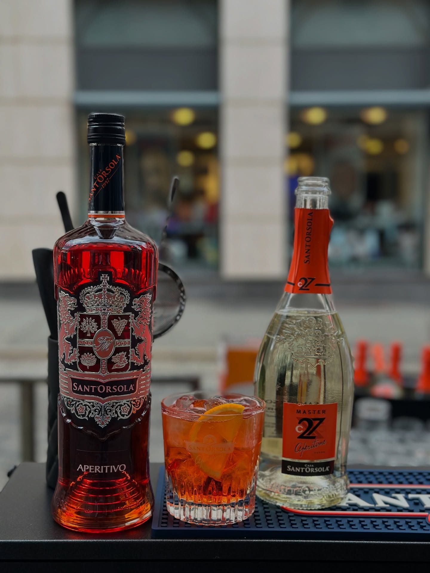 Due bottiglie di liquore rosso e una bevanda all'arancia su un tavolo da bar all'aperto.
