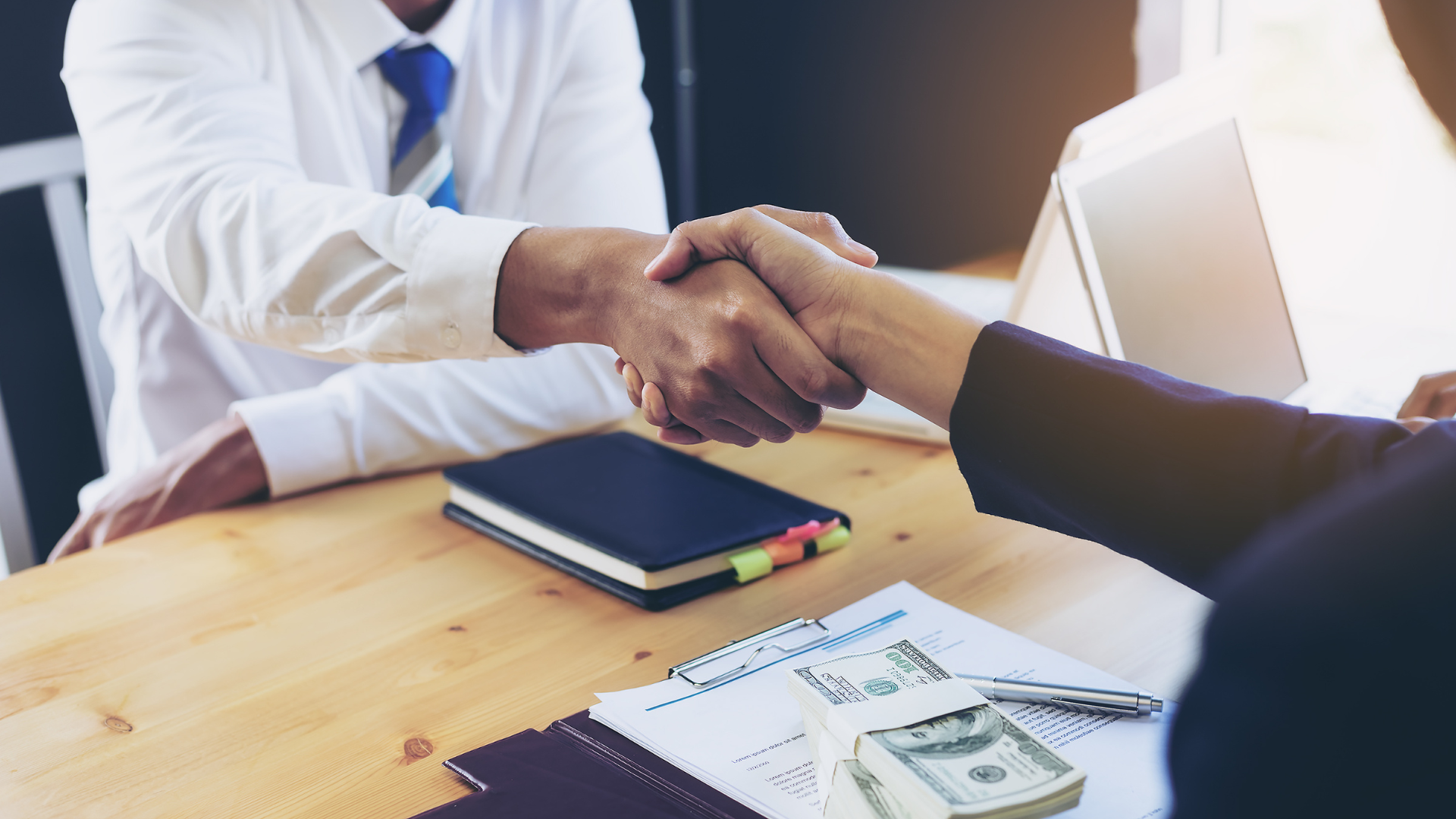 Two people in business attire shaking hands across a desk with documents and cash visible