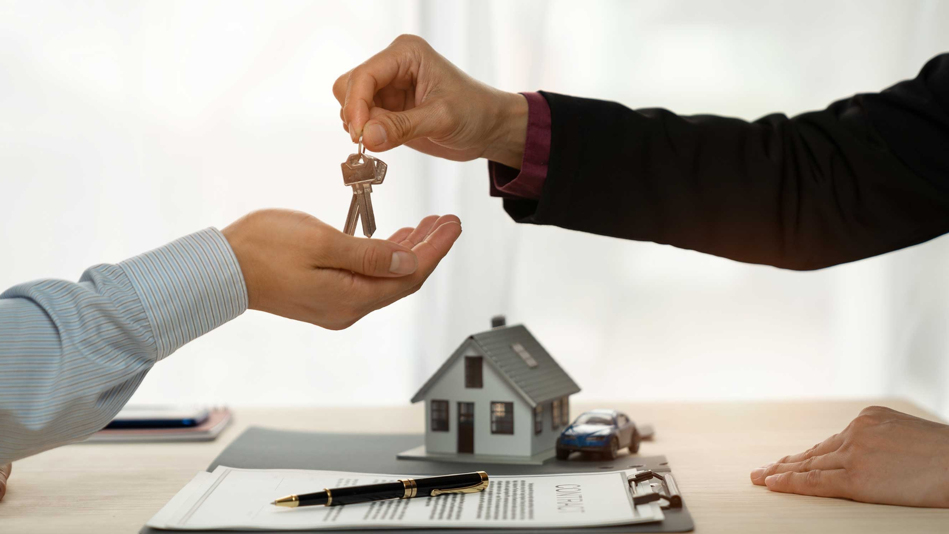 Hands exchange house keys over a model house and papers, suggesting a real estate transaction.