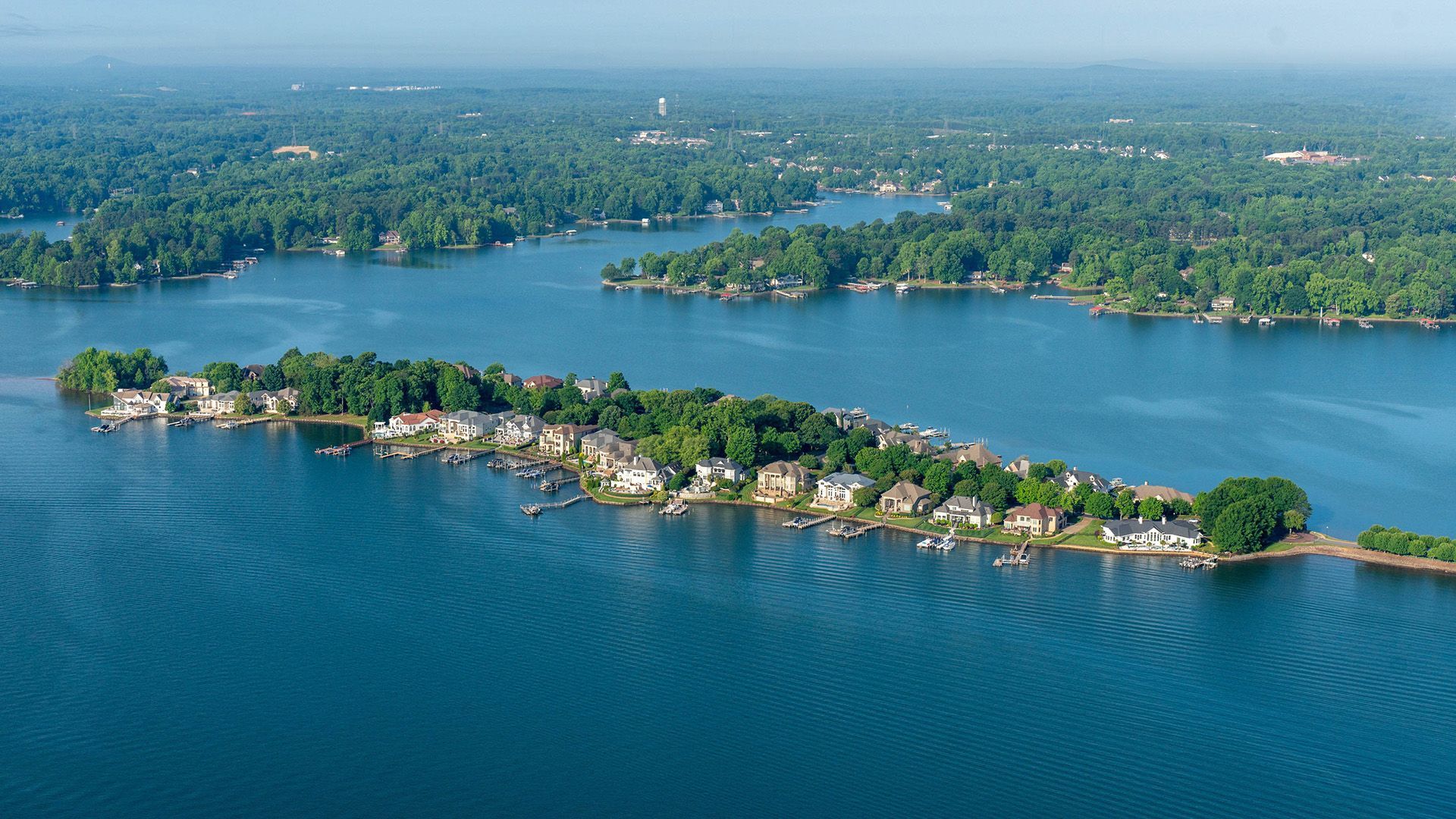 Aerial view of a wooded peninsula with homes, surrounded by calm blue water and distant shoreline.