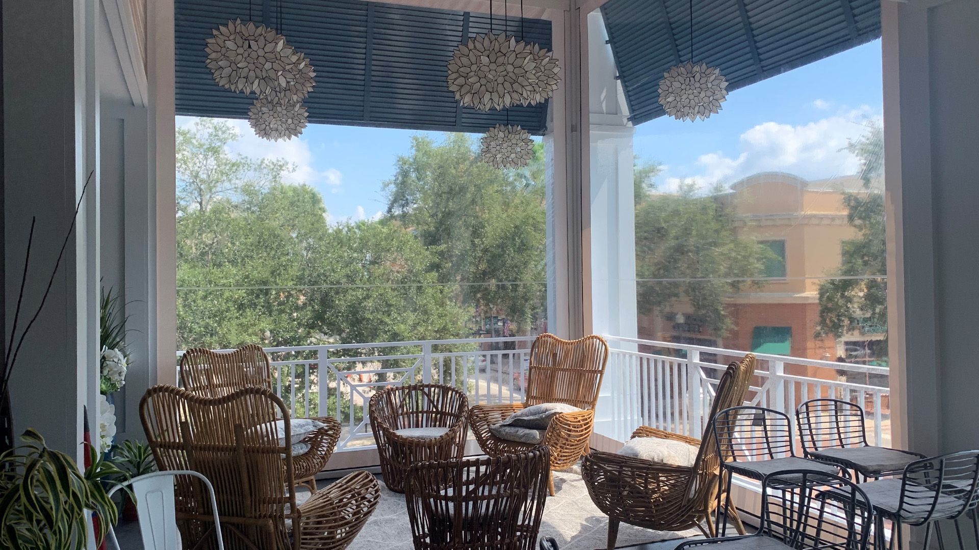 Outdoor café patio with wicker chairs and tables under a shaded awning, overlooking trees and a brick building.