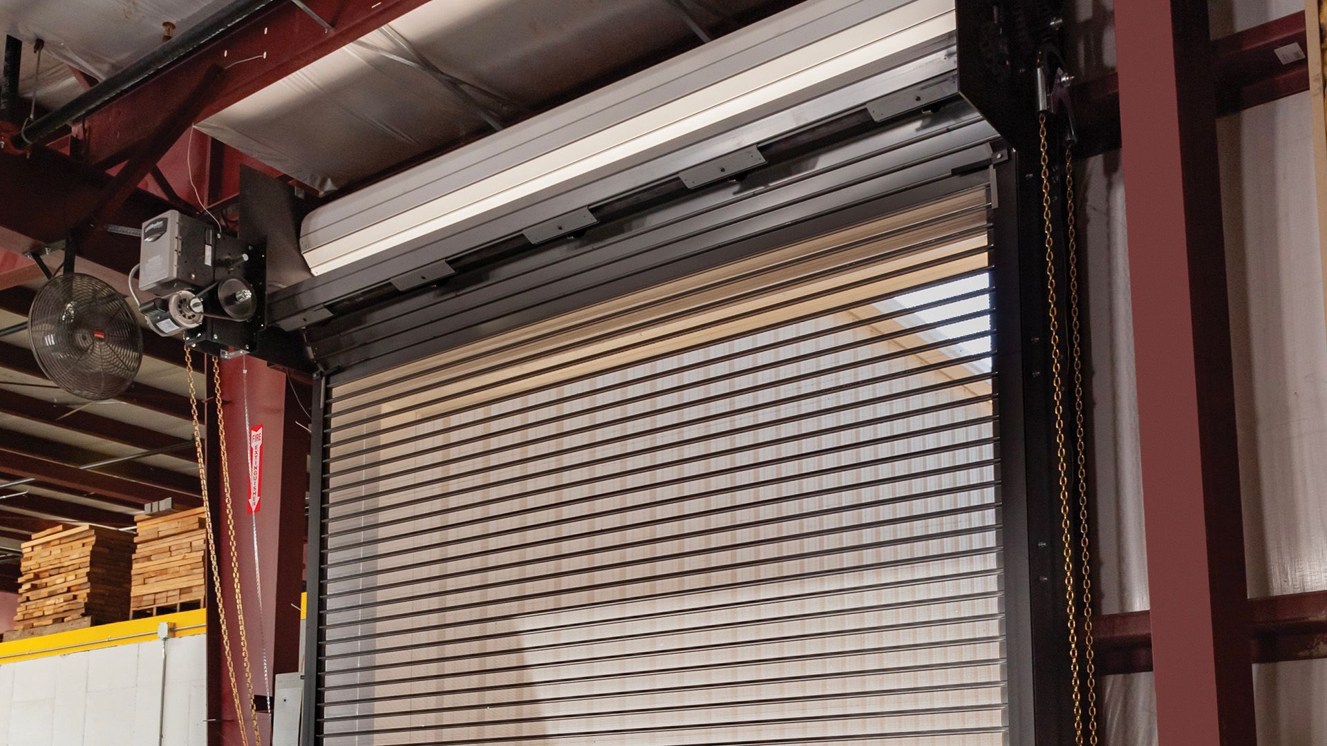 Industrial rolling shutter door inside a warehouse, partially closed under red and white machinery.