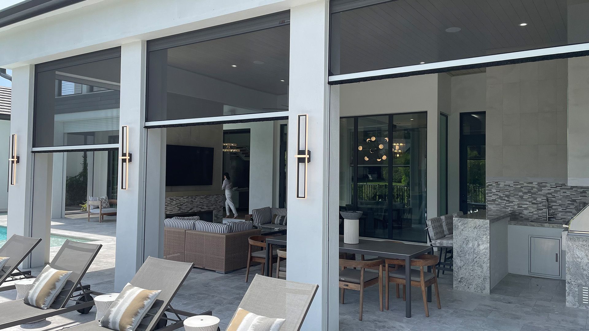 A luxury outdoor patio featuring retractable screens, a dining area, pool loungers, and a stone-built kitchen.