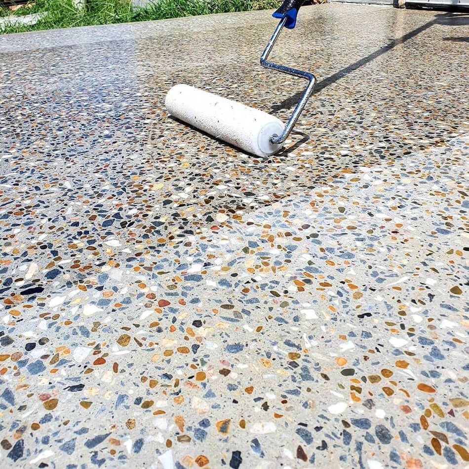 A paint roller applies a glossy sealant to a pebbled, exposed aggregate concrete surface outdoors.