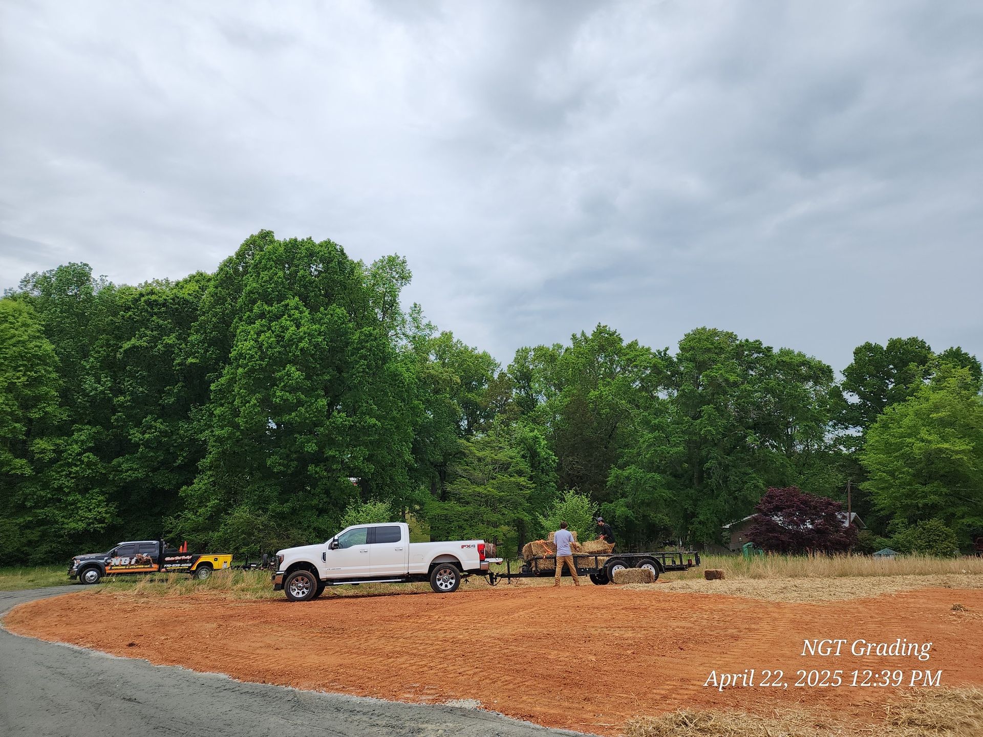 White pickup and other trucks parked on a red dirt lot beside trees under cloudy sky