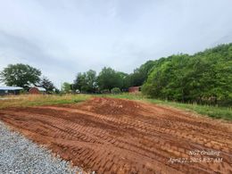 Freshly graded red dirt field beside green trees under a cloudy sky.