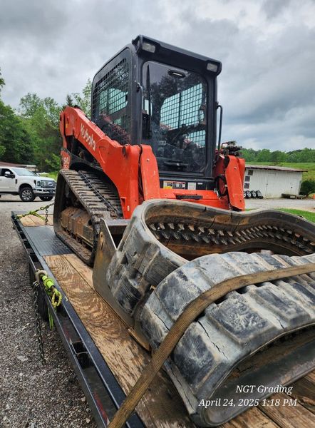 Orange skid-steer loader strapped on a flatbed trailer, viewed from the rear on a cloudy day.
