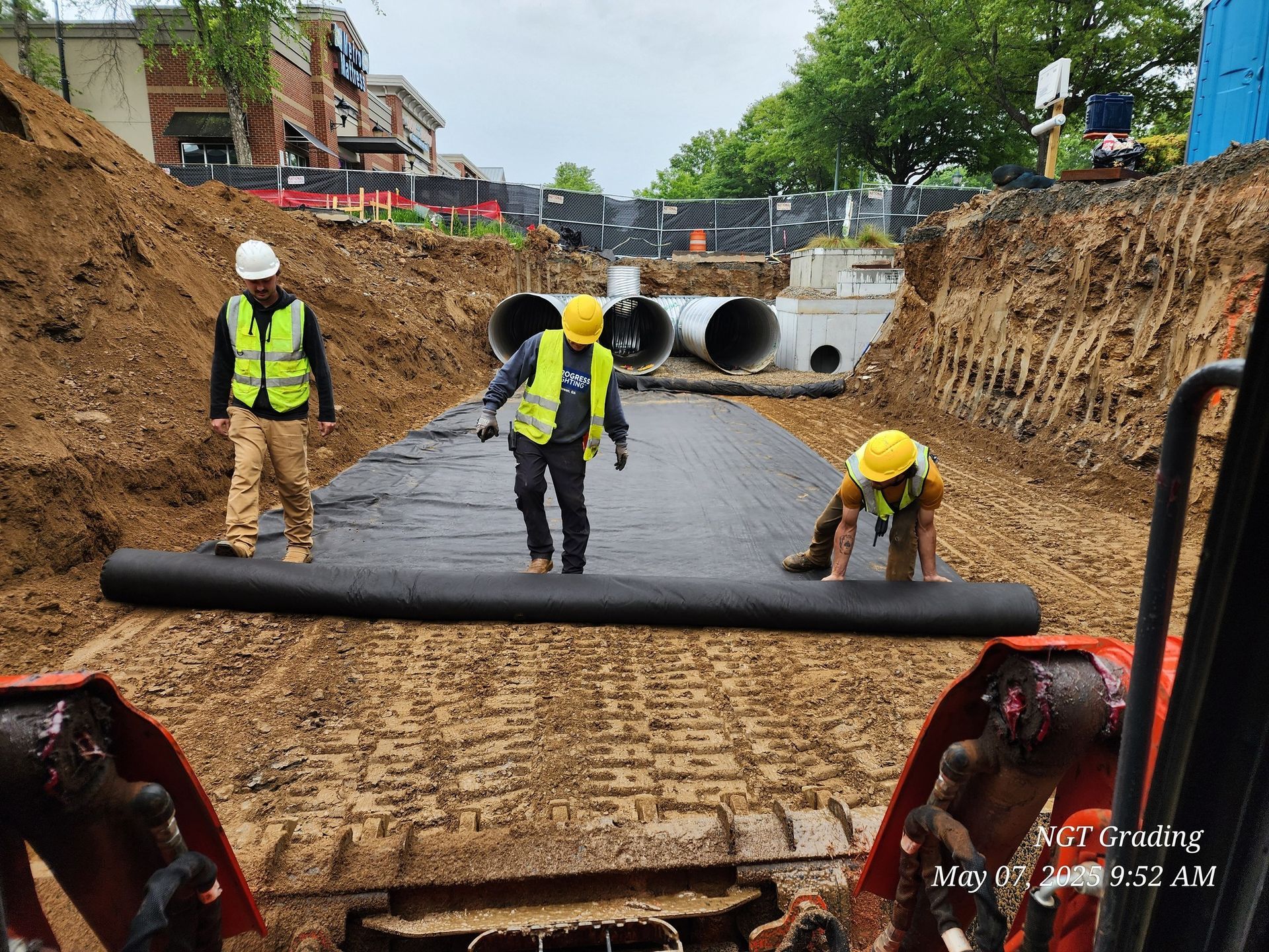 Construction workers laying black waterproof membrane in a trench with large concrete pipes ahead.