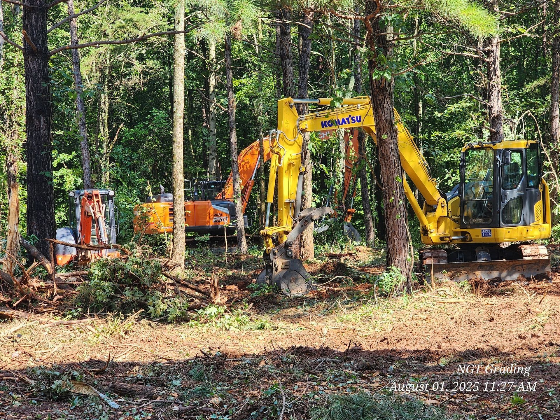 Yellow excavators and a truck clearing brush in a wooded forest area