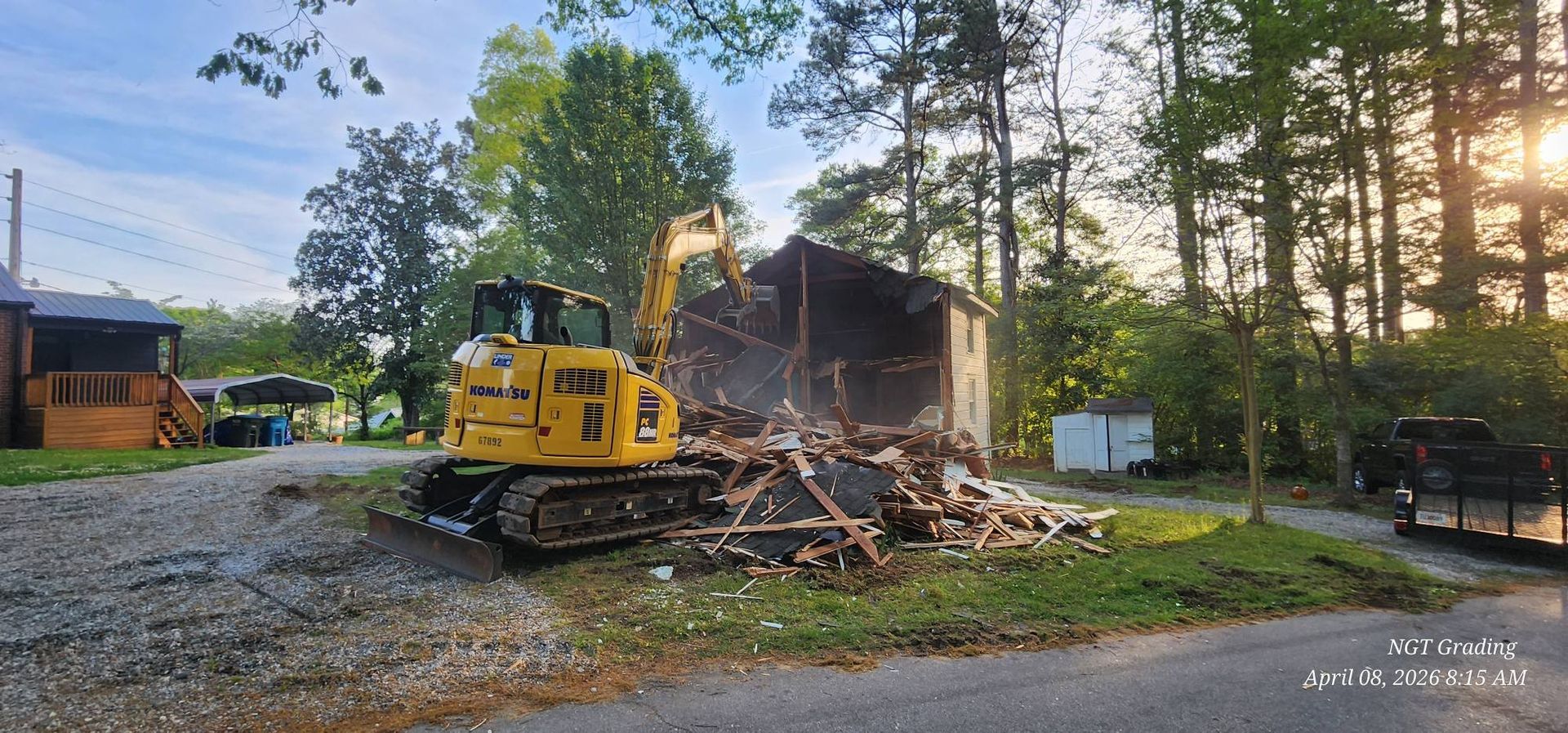 Yellow excavator demolishing a small house at dusk, with debris piled beside the road and trees nearby
