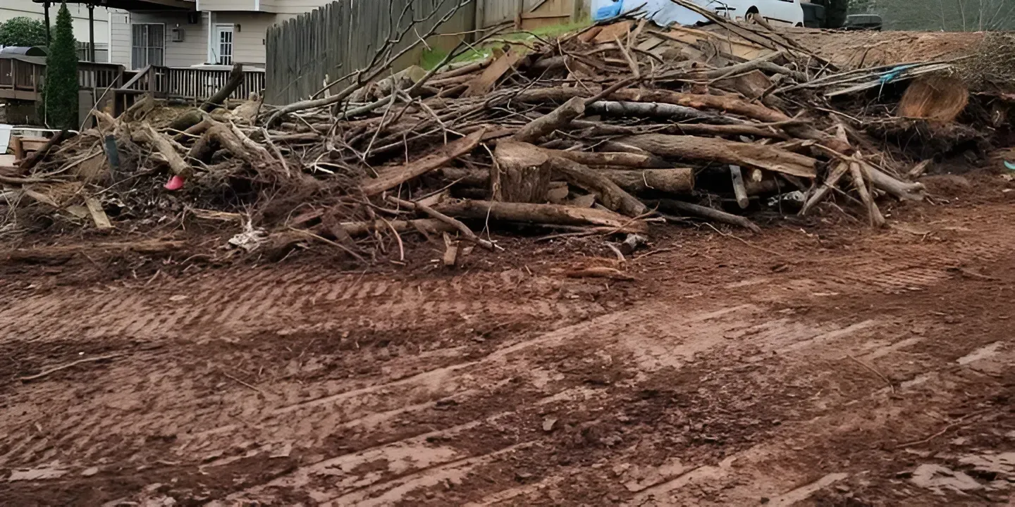 Pile of tree branches and logs beside a muddy dirt lot near a fence and house