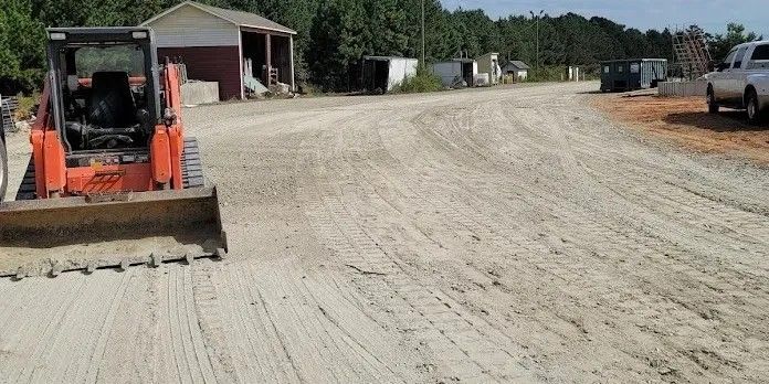 Orange skid steer grading a gravel driveway near a building and parked truck.