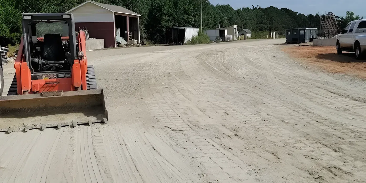 Orange skid steer grading a gravel road near a small building and parked trucks.