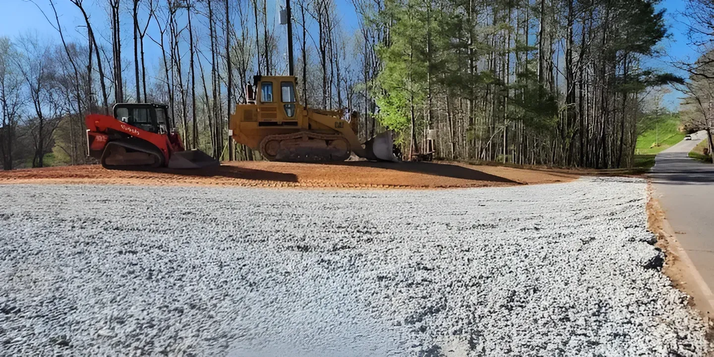 Construction vehicles grading a roadside dirt lot beside a paved street and trees