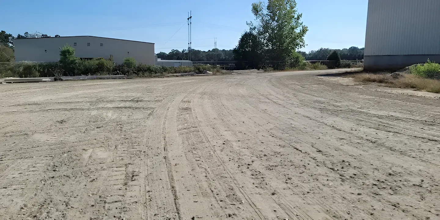 Empty dirt lot with tire tracks, flanked by two white buildings and trees under a clear sky