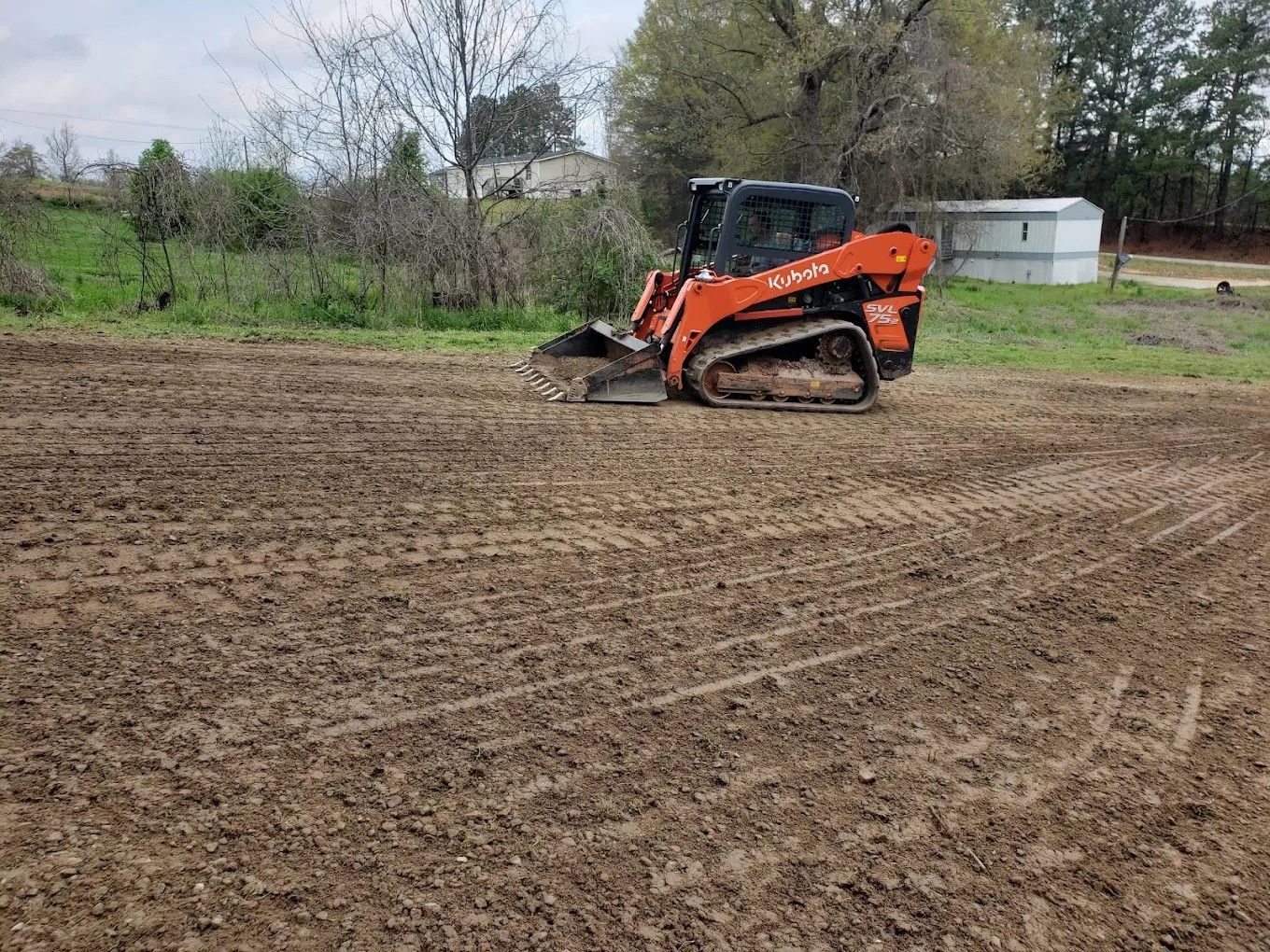 Orange skid-steer loader grading a muddy field near trees and a small white shed.