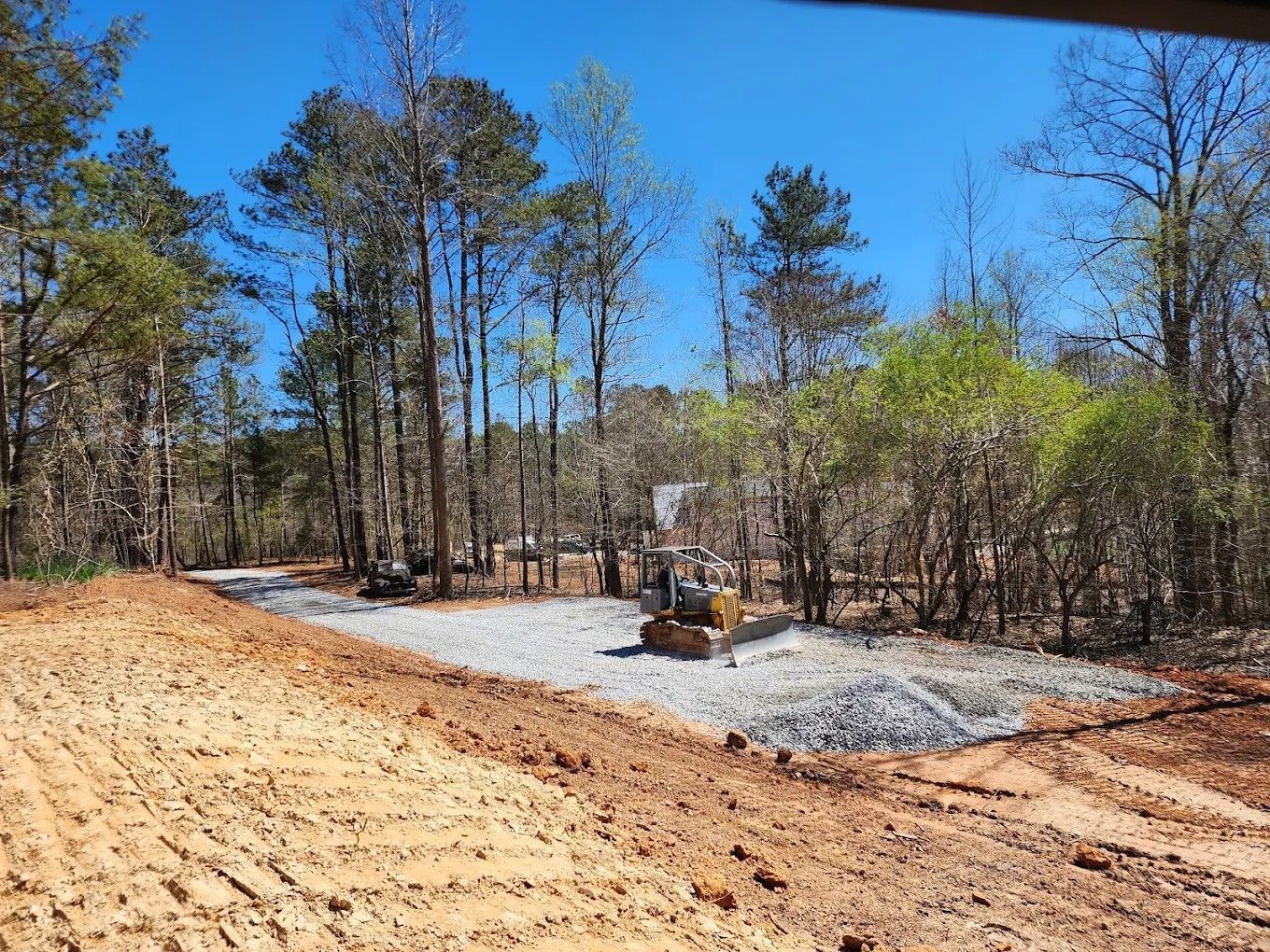 Gravel driveway under construction beside a wooded lot, with a bulldozer and cleared earth under a blue sky.