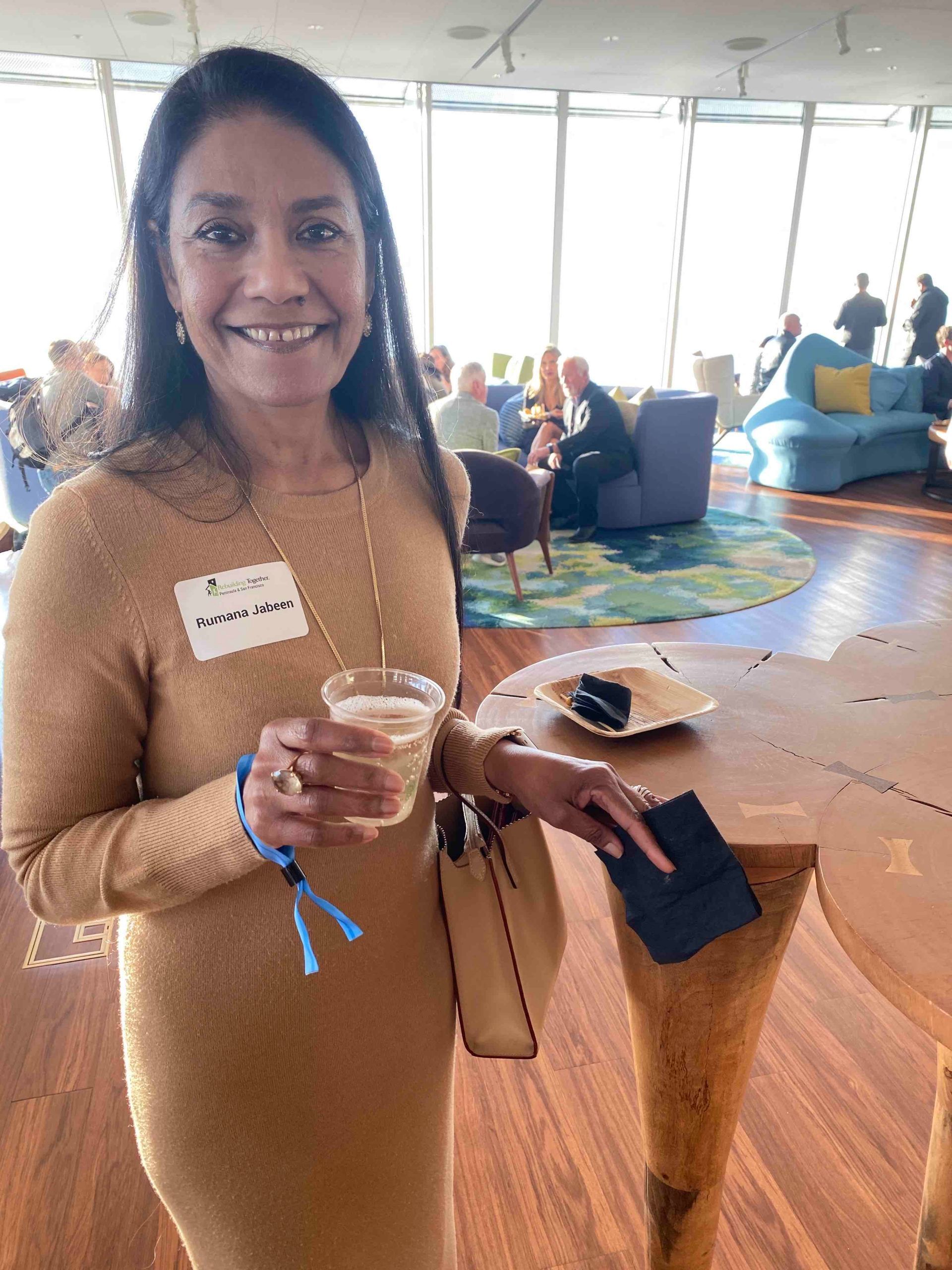 A woman in a tan dress is holding a drink and smiling.