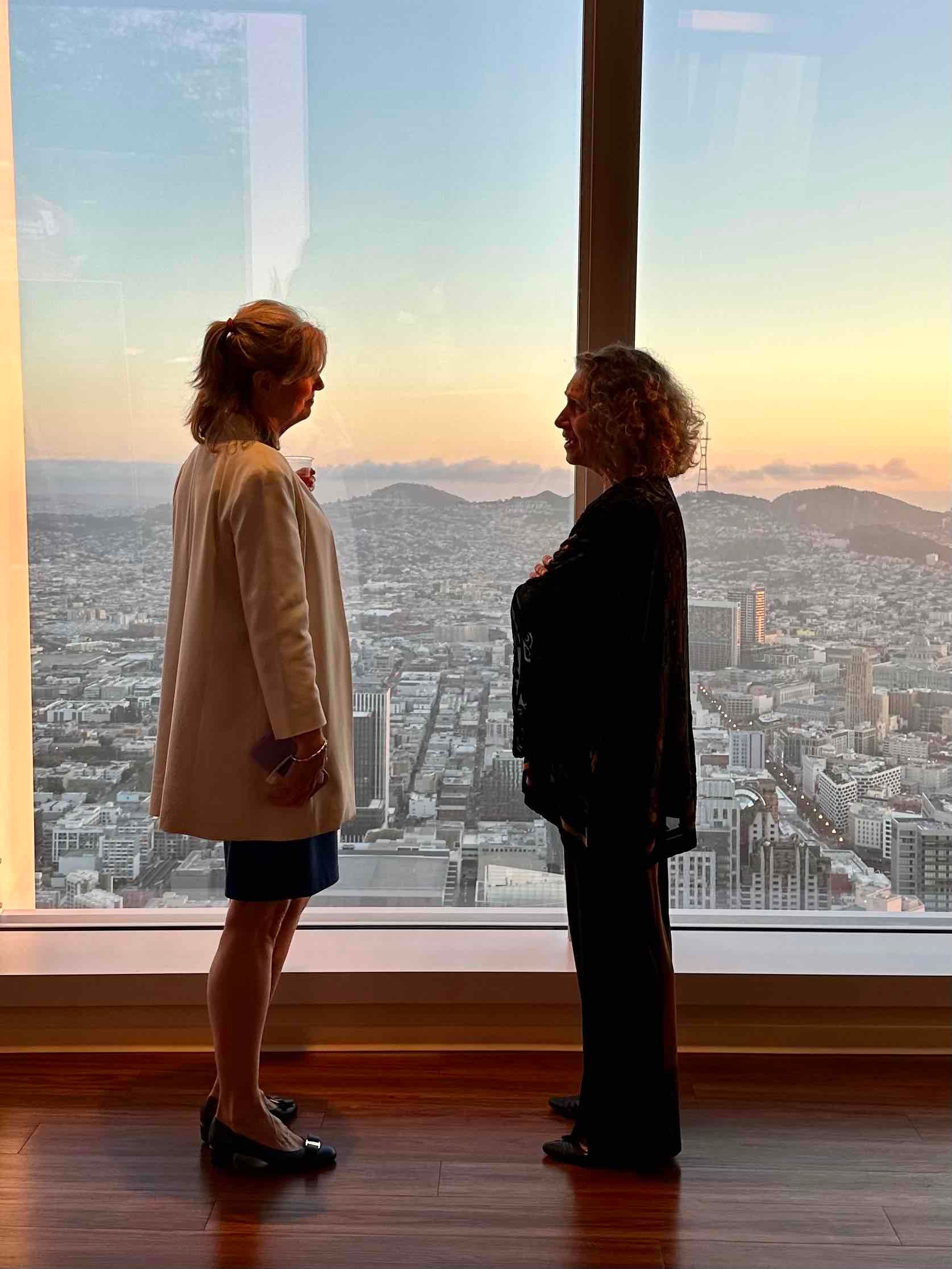 Two women are standing next to each other in front of a window overlooking a city.