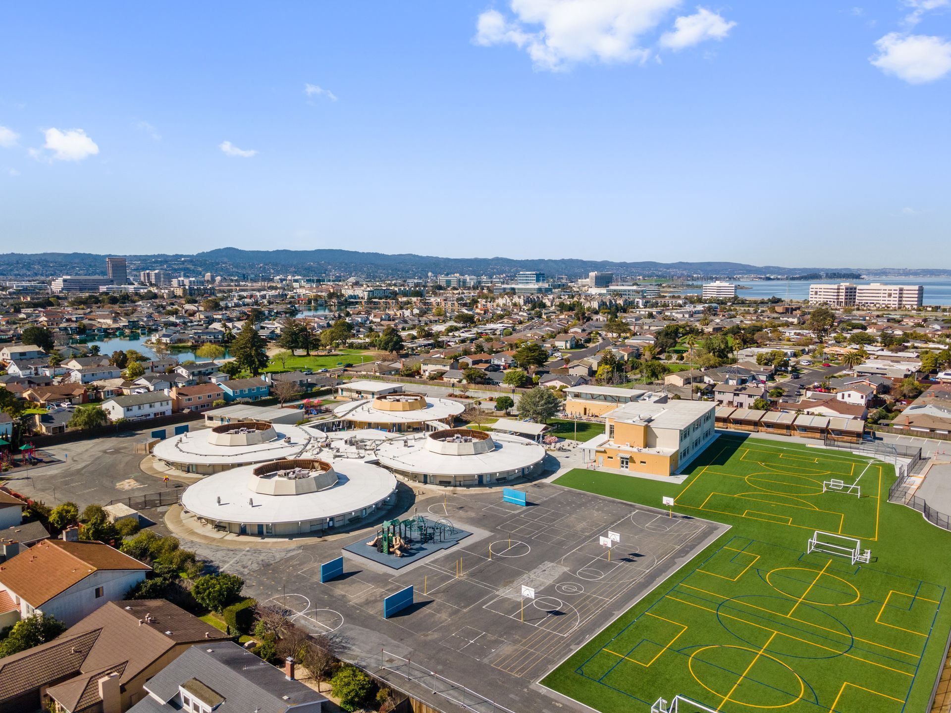 An aerial view of a school with a soccer field in the foreground and a city in the background.
