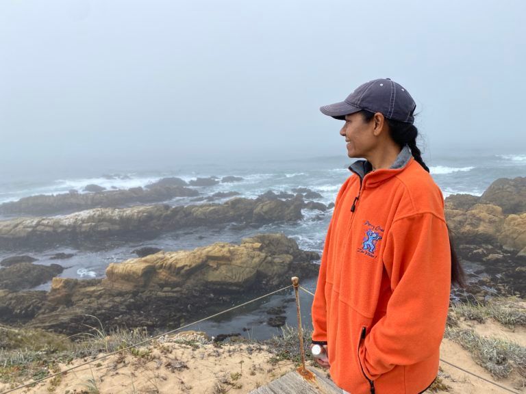 A woman in an orange jacket and hat stands on a rocky cliff overlooking the ocean