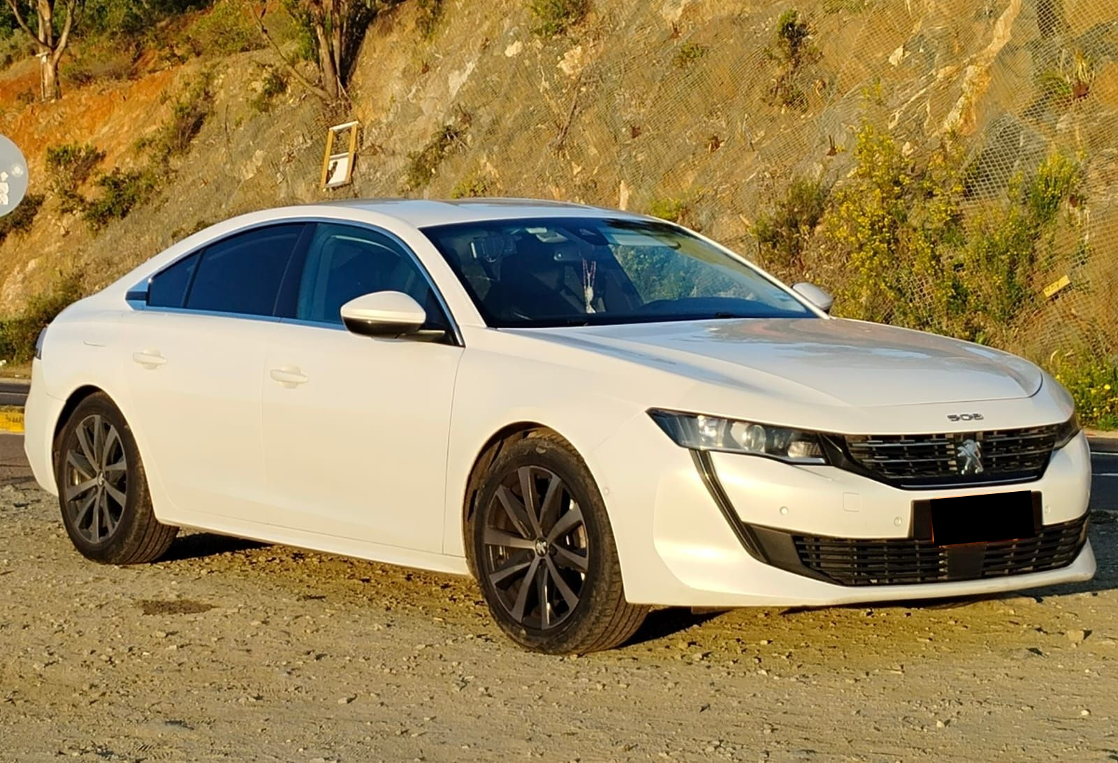 Un sedán Peugeot 508 blanco estacionado sobre una superficie de grava con una ladera rocosa al fondo.