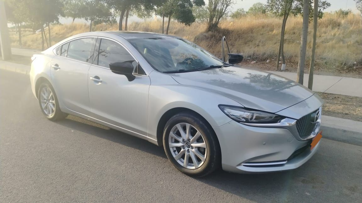 Un sedán Mazda 6 plateado estacionado al costado de una carretera pavimentada, con árboles y hierba seca al fondo.