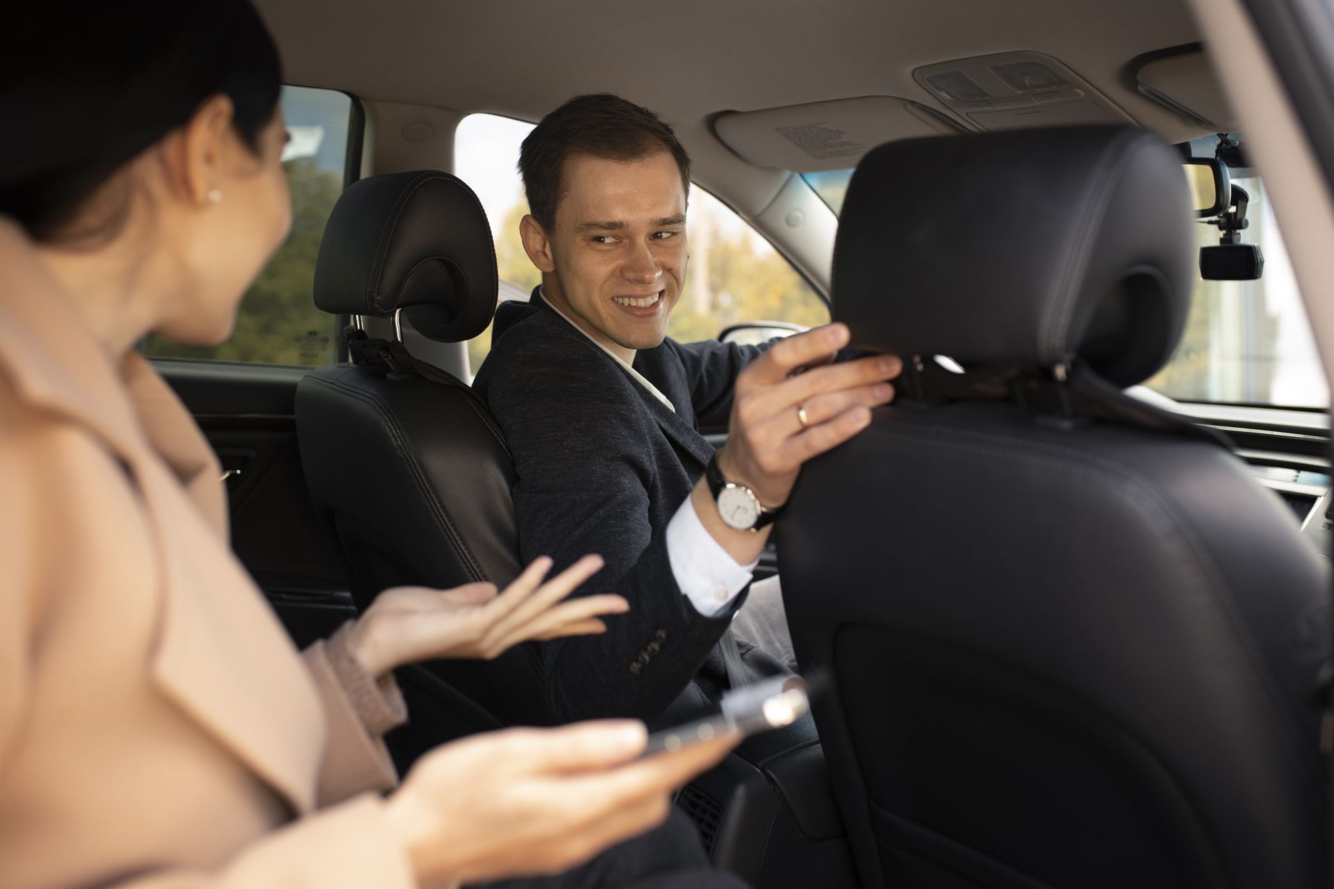 Un hombre de traje sonríe, ajustando un reposacabezas mientras una mujer sostiene el teléfono en el automóvil.