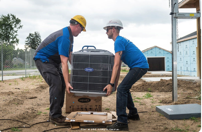 Two construction workers in blue shirts and hard hats lift a large air conditioning unit outdoors.