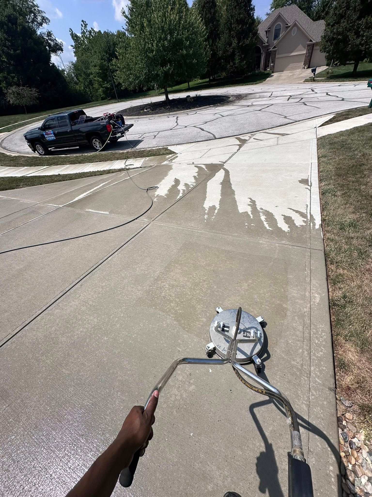 Concrete driveway being pressure washed, with a spray wand in the foreground and a car parked nearby.