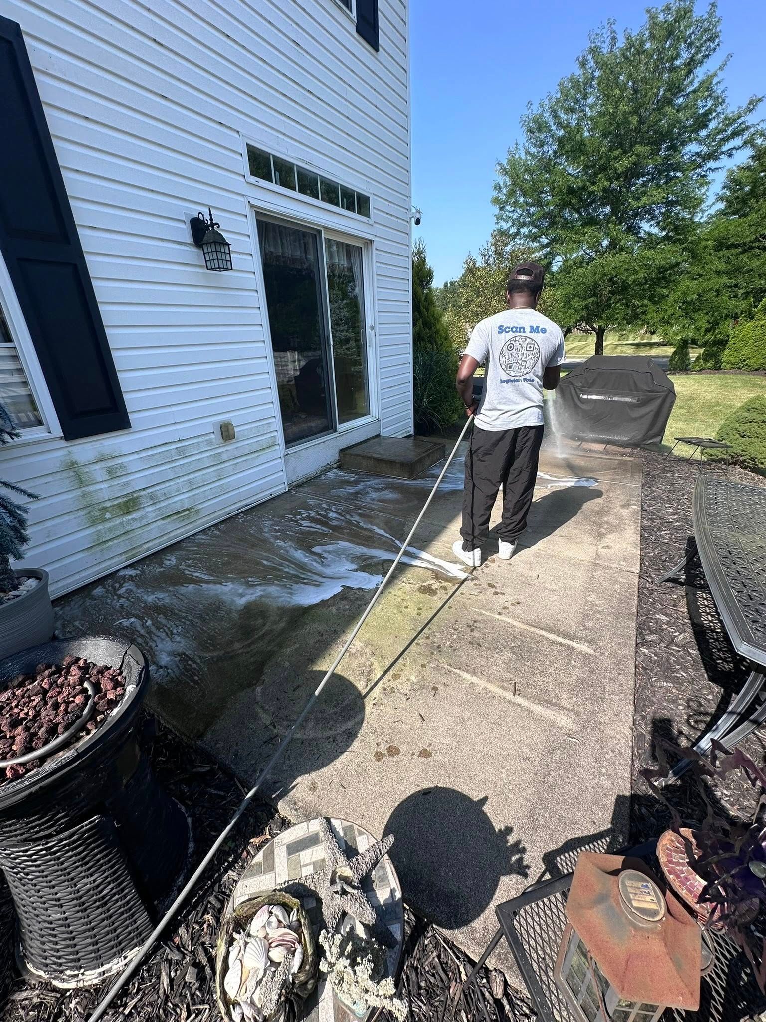 Person pressure-washing a house walkway beside white siding and potted plants