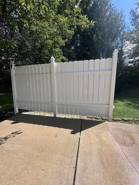 White vinyl privacy fence across a sunlit concrete driveway, with trees in the background.