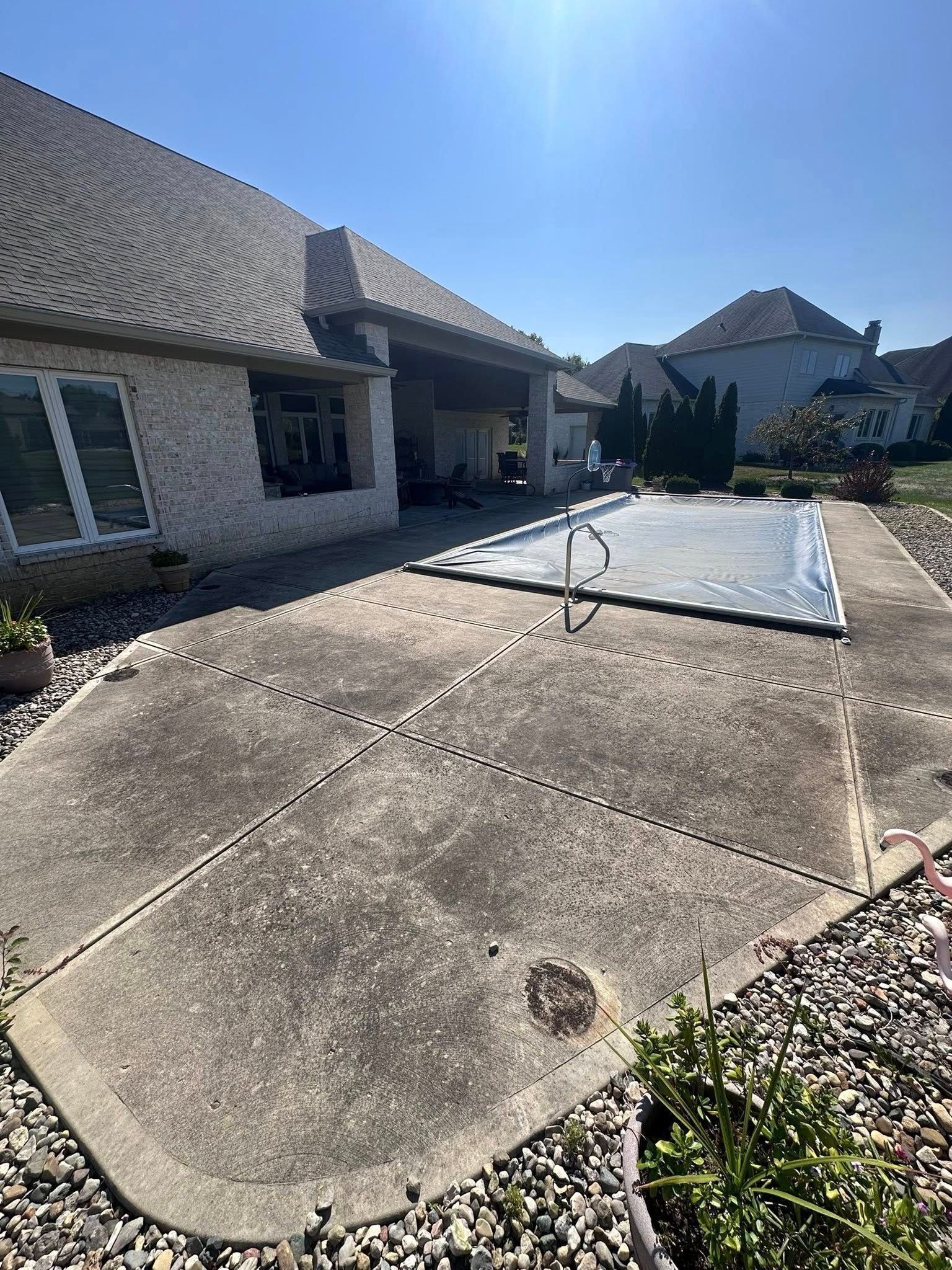 Concrete driveway and patio beside a stone house under a bright blue sky, with a rectangular board covering part of the drive