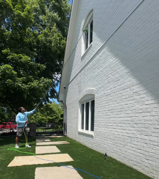 Person spraying the side of a white brick house in a sunny yard, with a ladder nearby