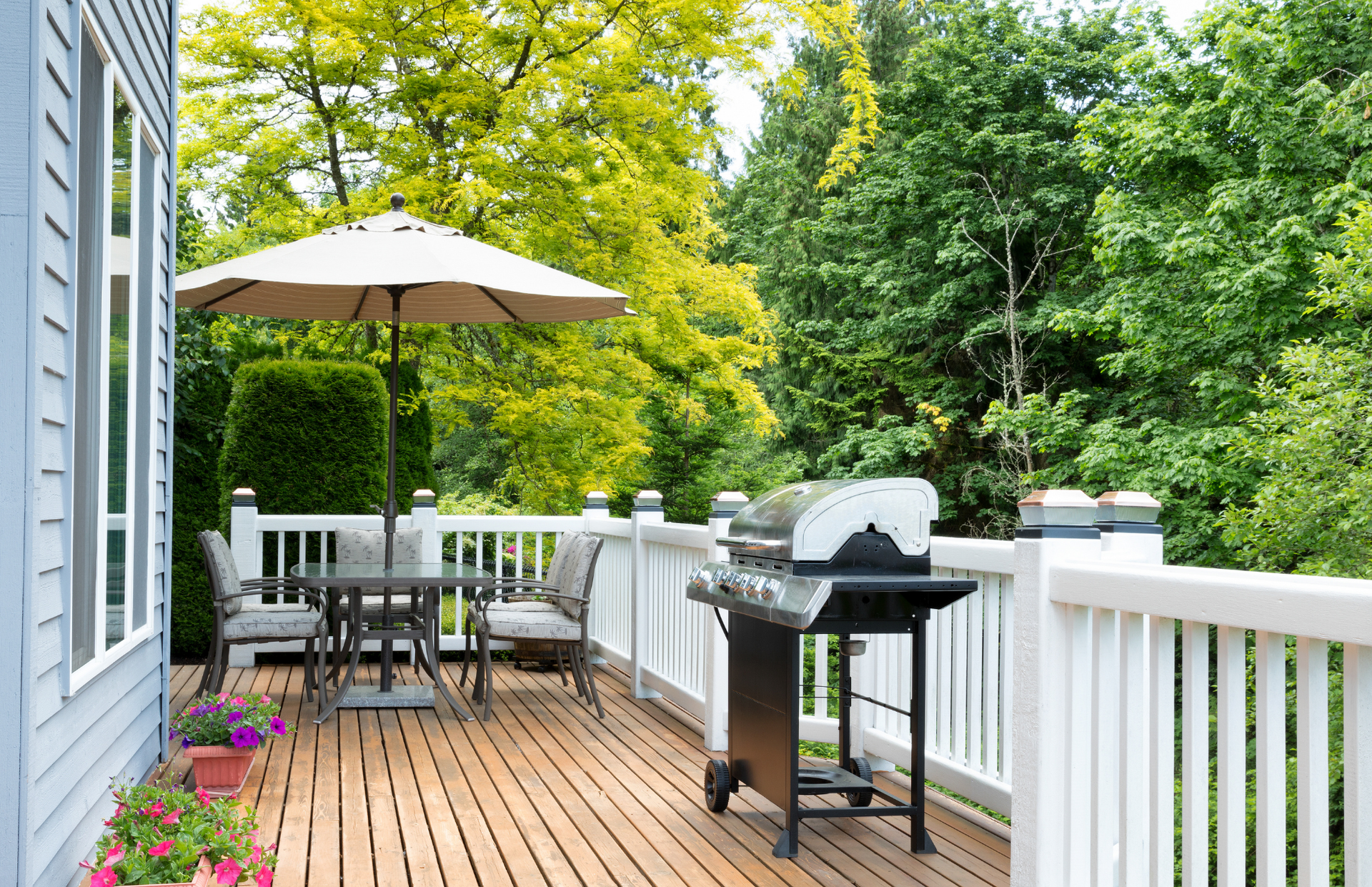 Wooden deck patio with table, umbrella, grill, and white railing surrounded by green trees
