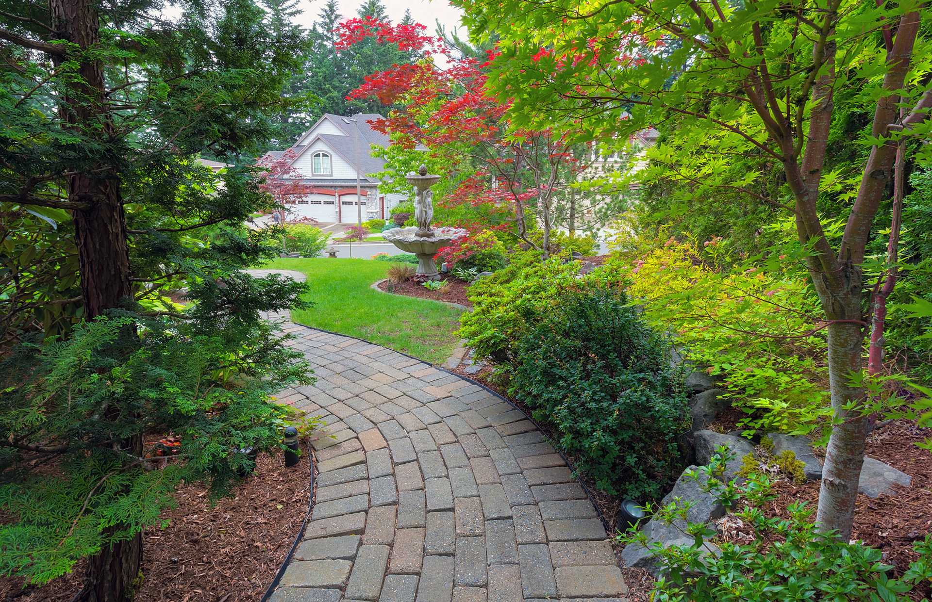 Curved brick garden path leading through lush greenery to a white house with red roof in the distance