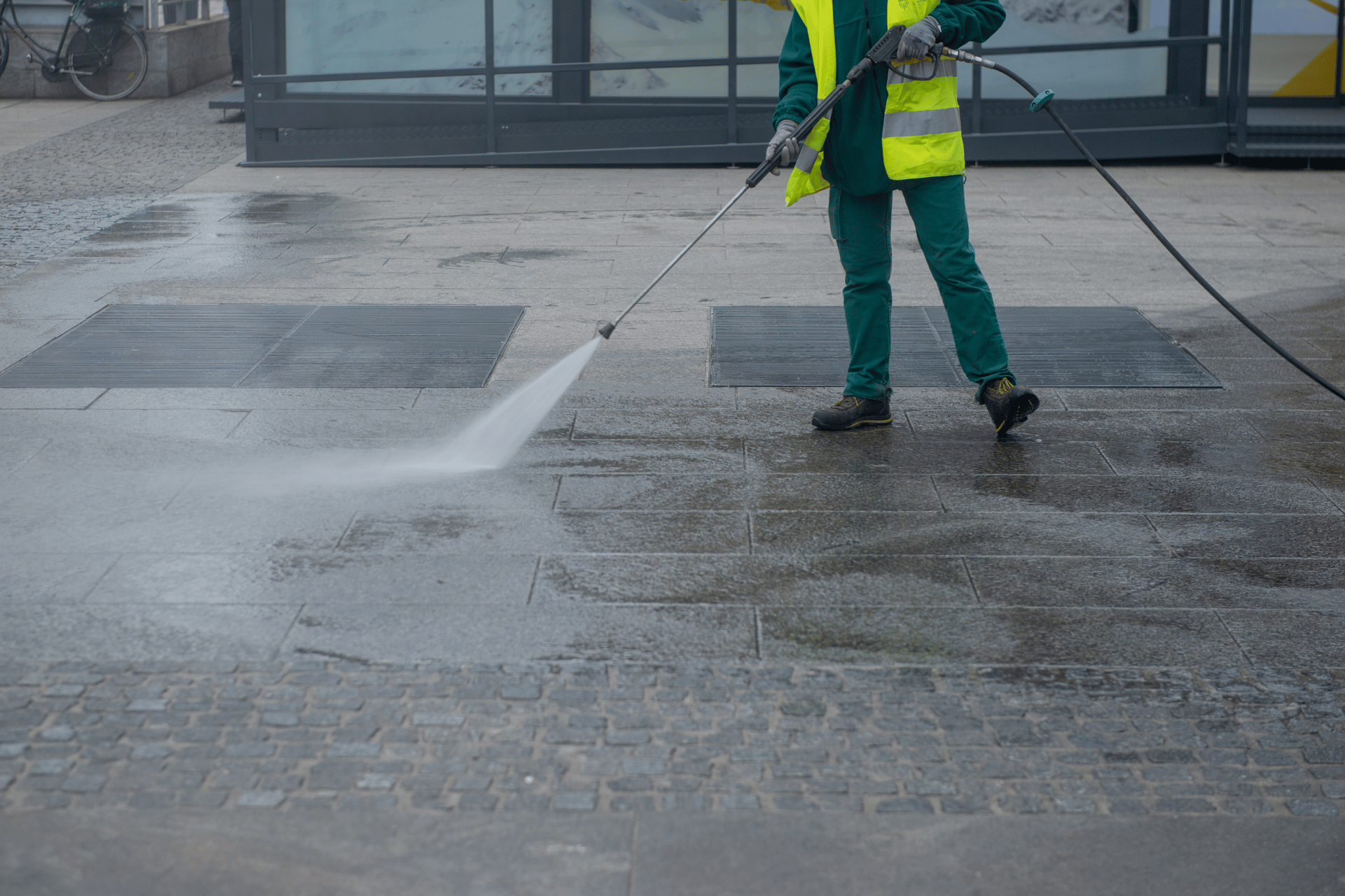 Worker in a yellow vest power-washing a wet sidewalk outside a building