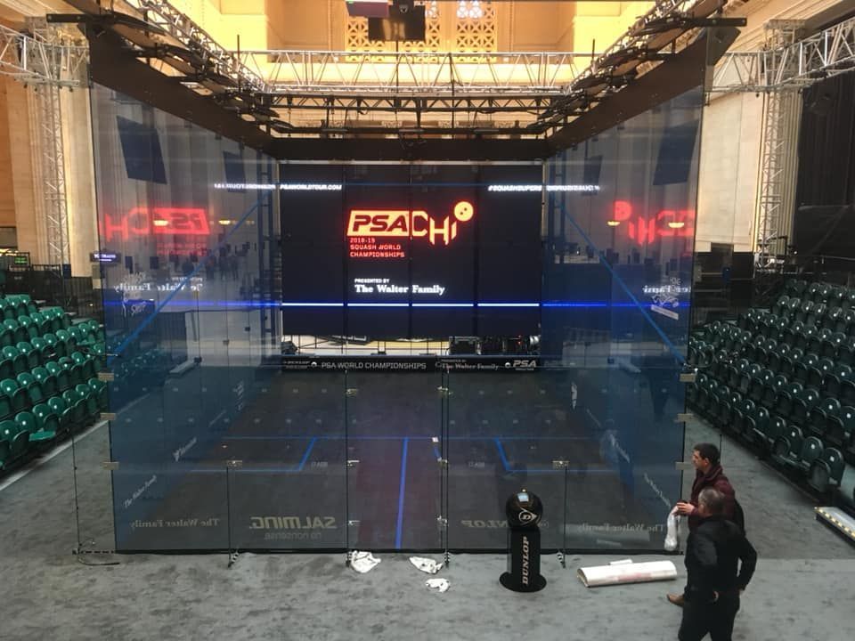 Indoor boxing ring with glass walls and “PSAH” banner, flanked by rows of green seats under stage lighting.
