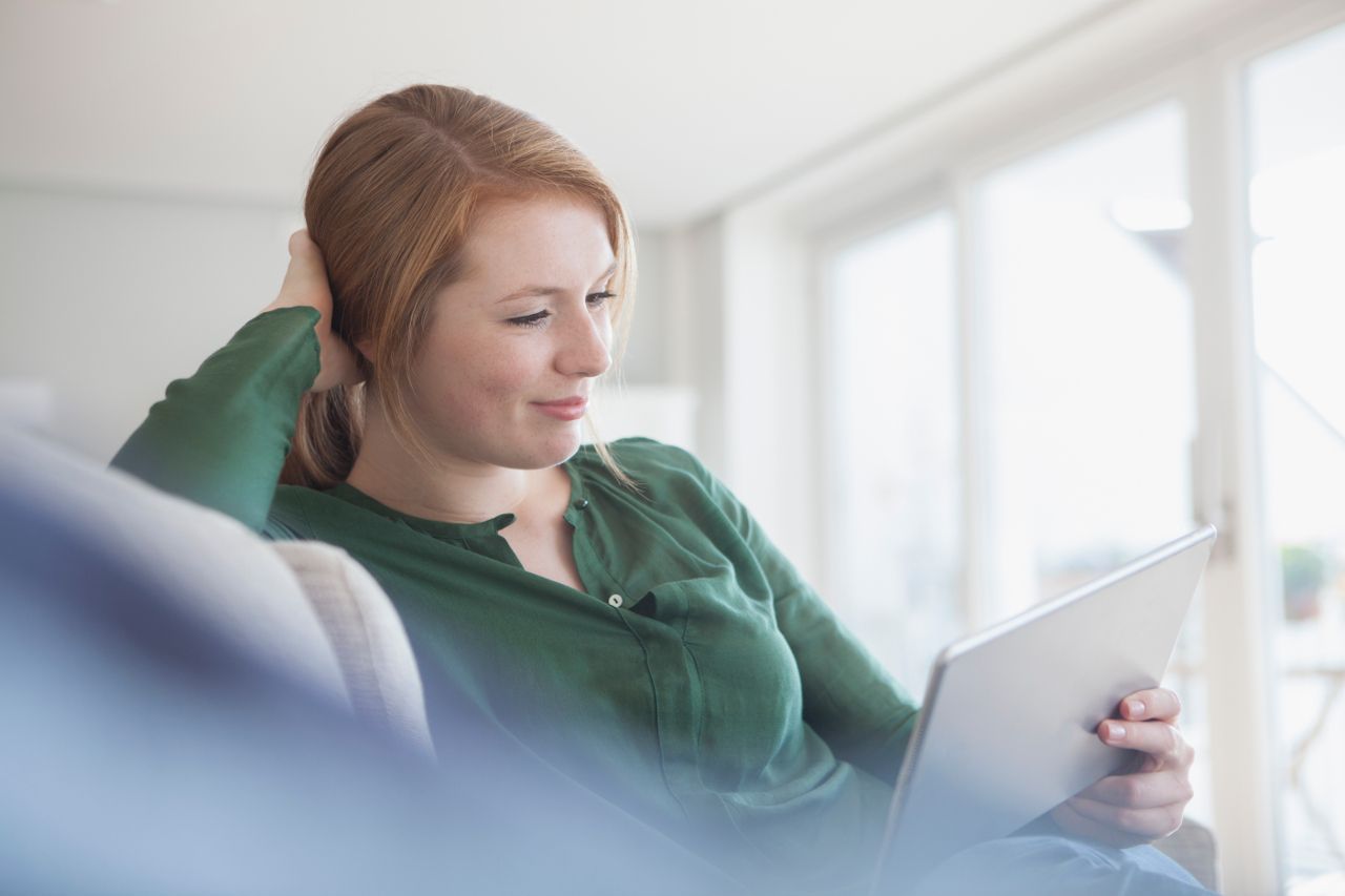 A person in a green shirt sits on a sofa, looking at a tablet with a gentle smile.