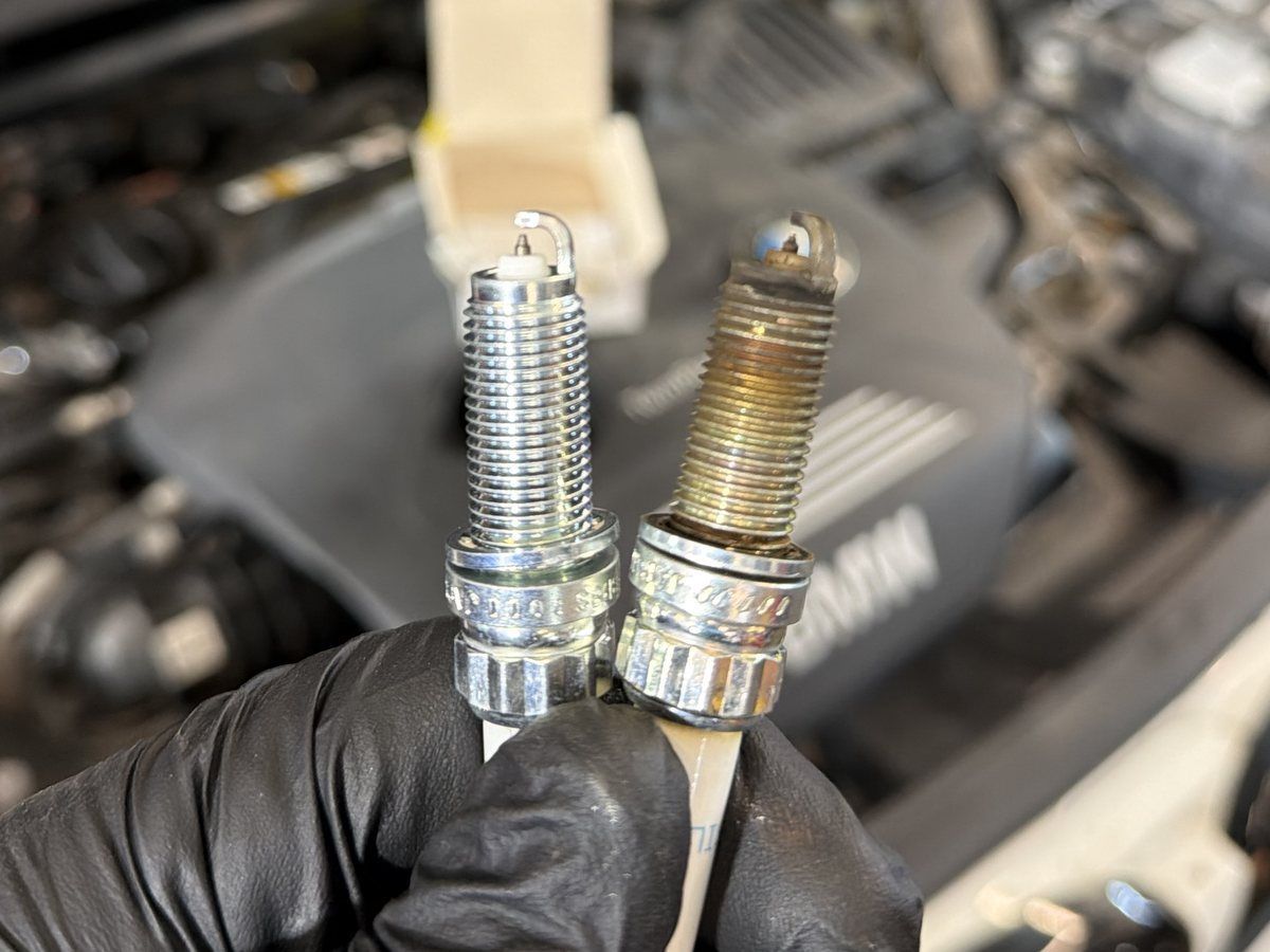 A new spark plug next to a worn one, held in front of an engine at No Name Auto Repair in Norwood, MA