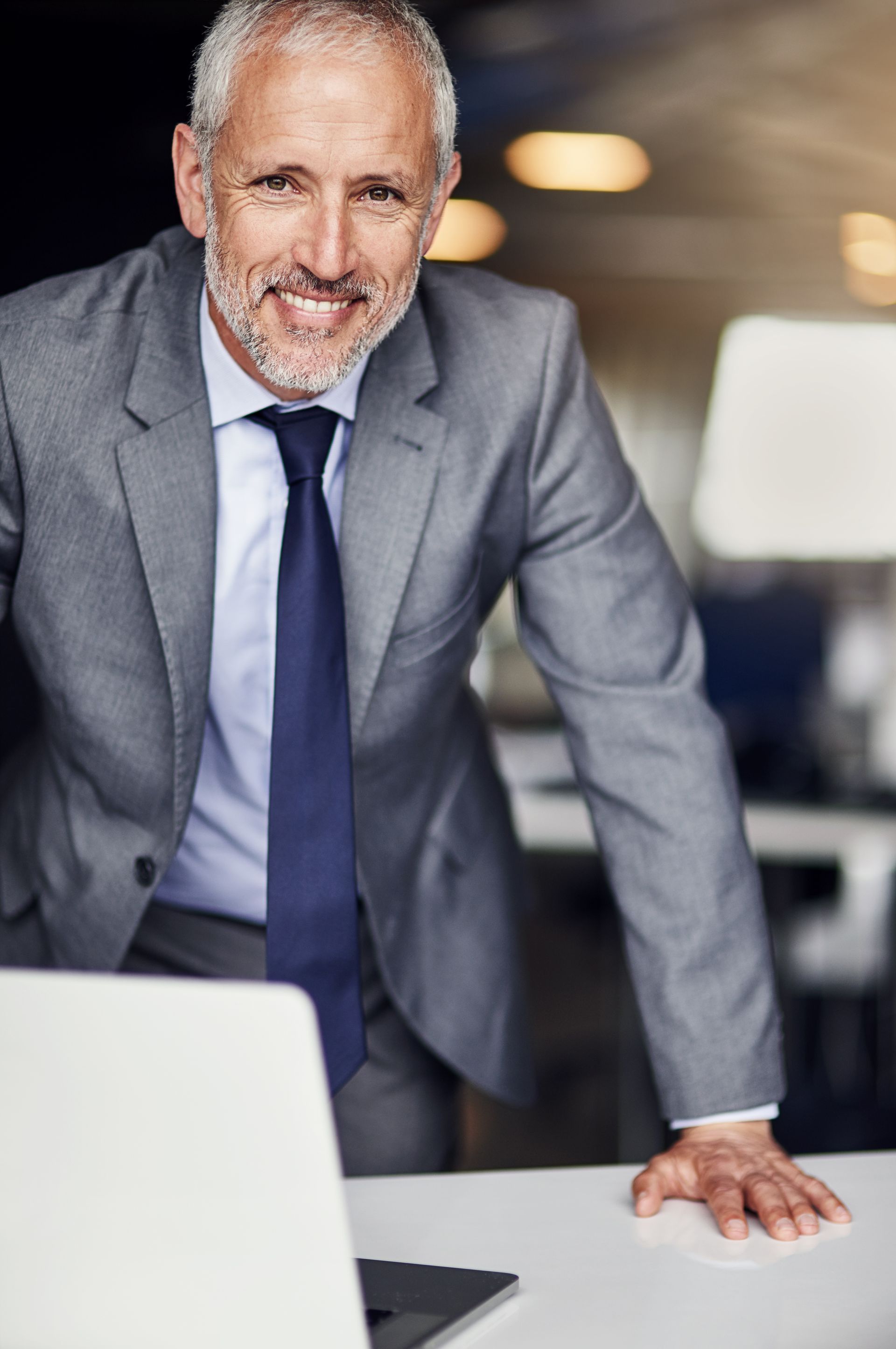 A man in a suit and tie is leaning on a desk in front of a laptop computer.
