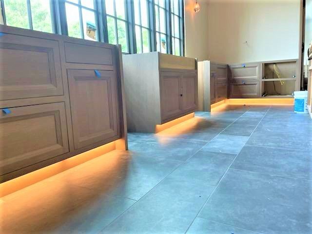 Modern kitchen with gray cabinets, large windows, and warm under-cabinet lighting along a tiled floor