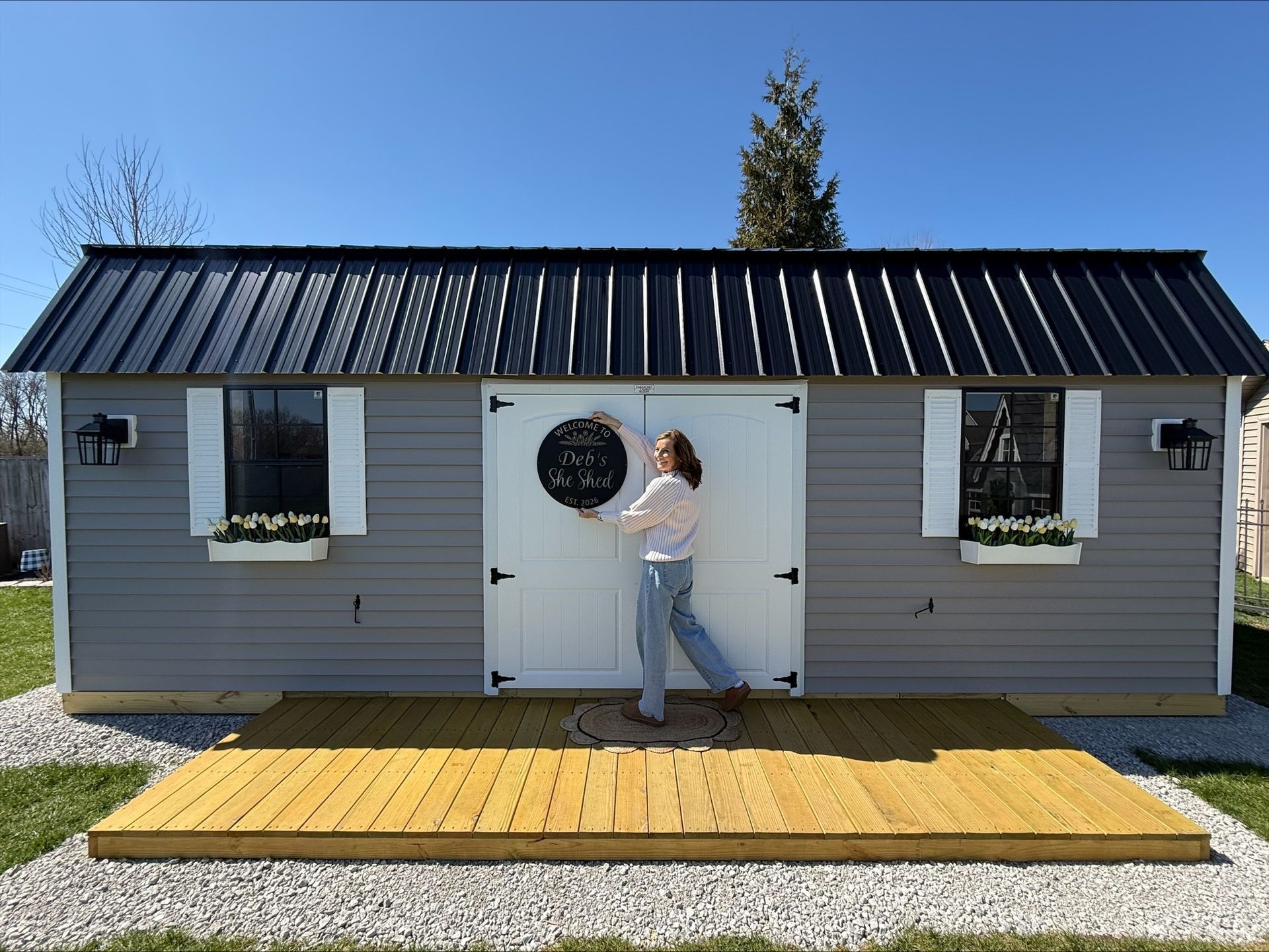 Small gray cabin with white doors and a person standing on a wooden deck under a blue sky