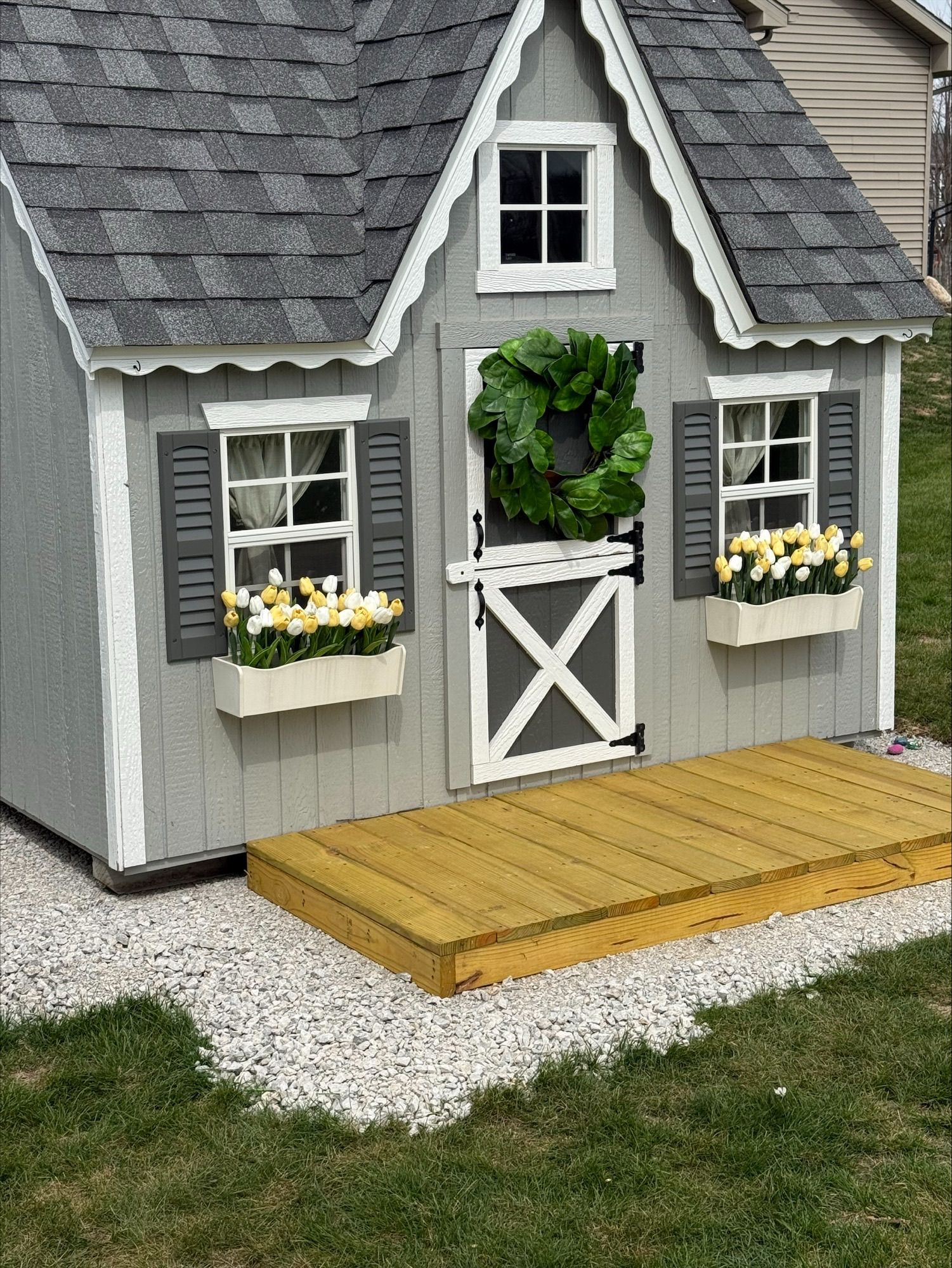 Small gray playhouse with white trim, flower boxes, and a wooden ramp on gravel.