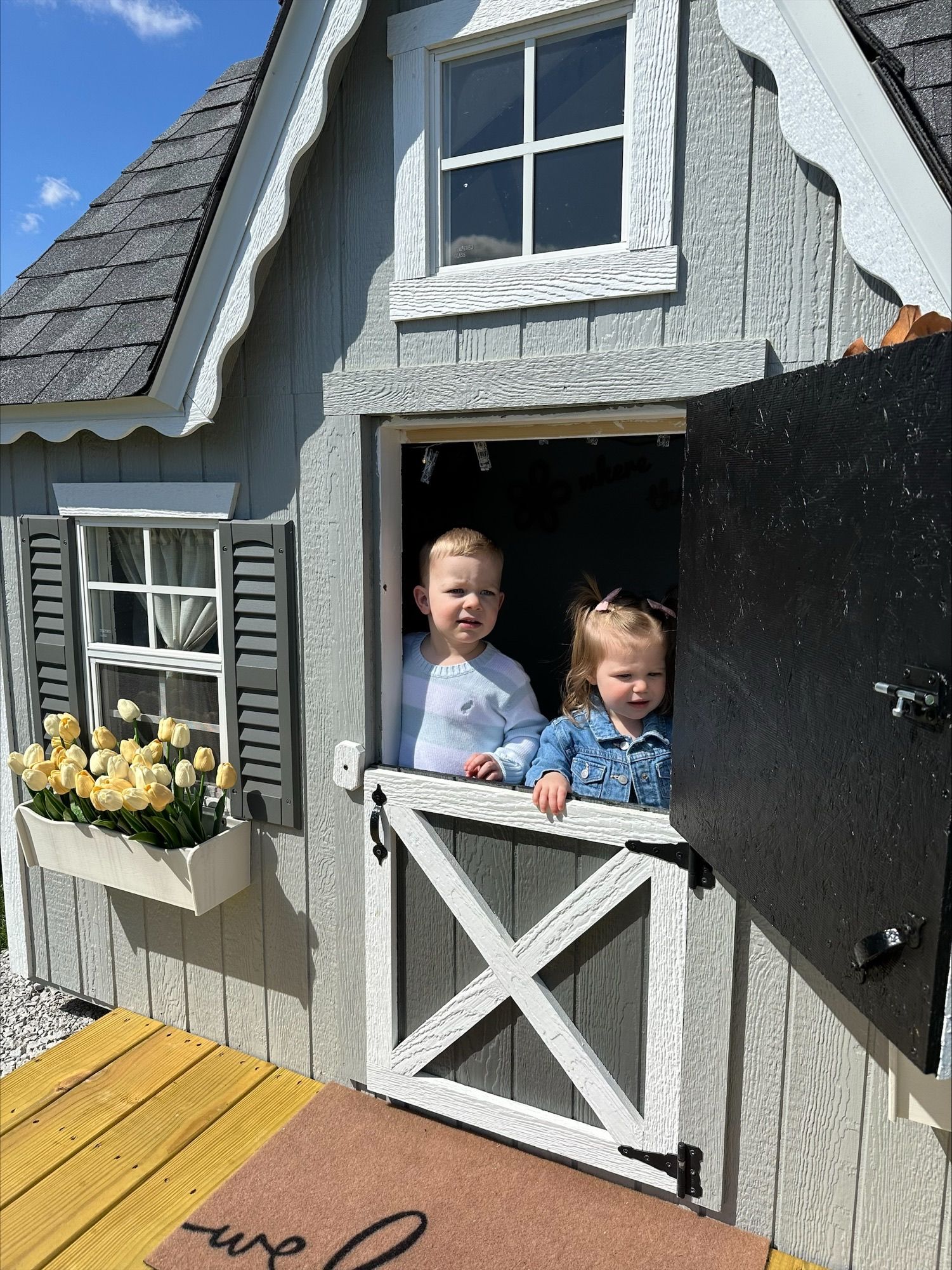 Two children smiling in a small playhouse window with a flower box and open black door