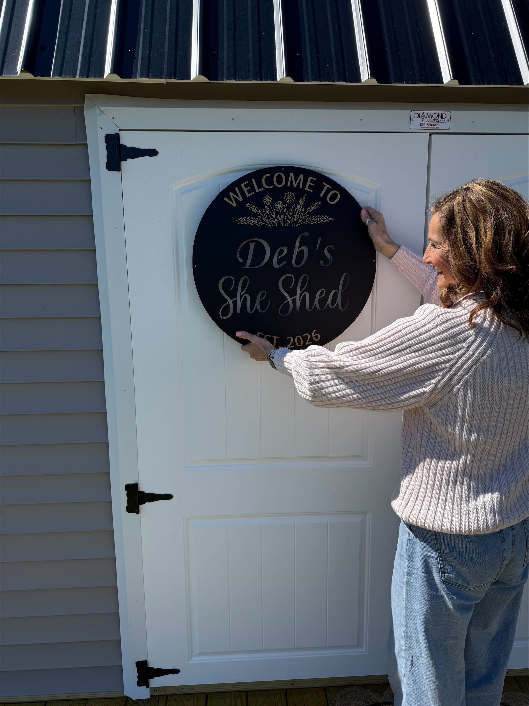 Woman hanging a round Welcome to Deb’s She Shed sign on a white door
