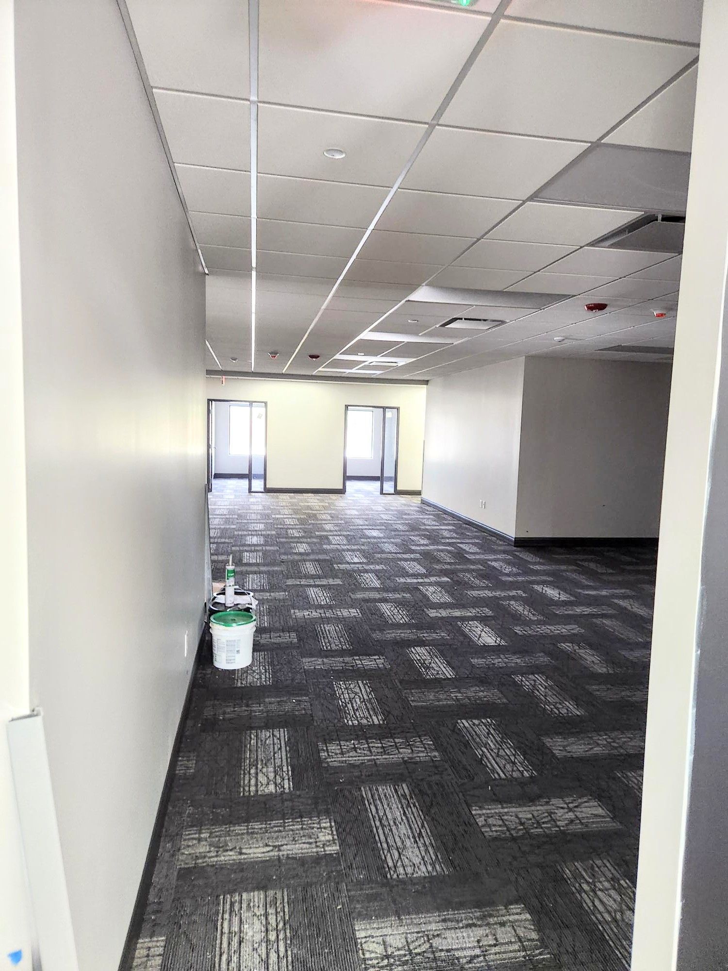 Empty office hallway with gray carpet, white walls, ceiling tiles, and a mop bucket near windows at the far end