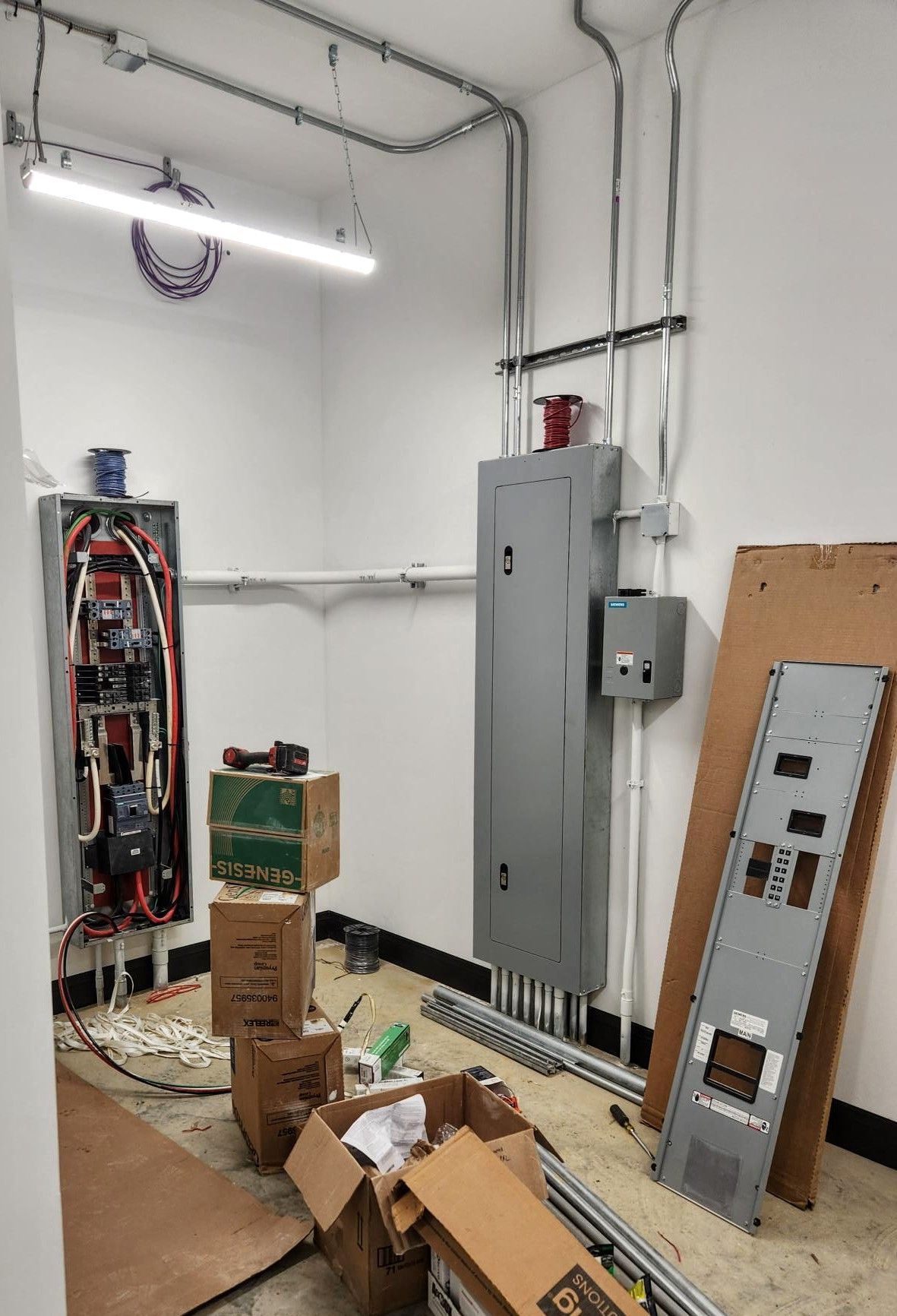 Electrical room with open panels, gray breaker cabinet, scattered boxes, and a detached metal panel on the floor.