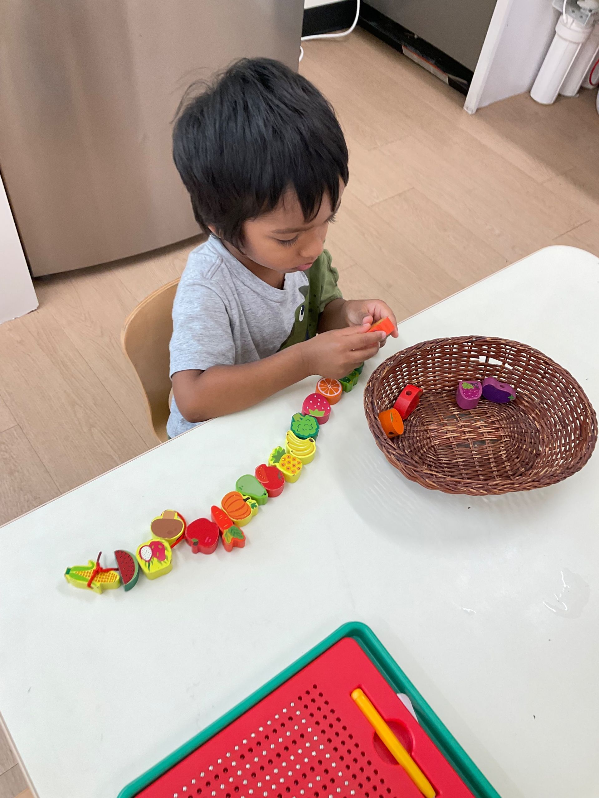 Child playing with colorful toy food on a table beside a brown basket in a bright room