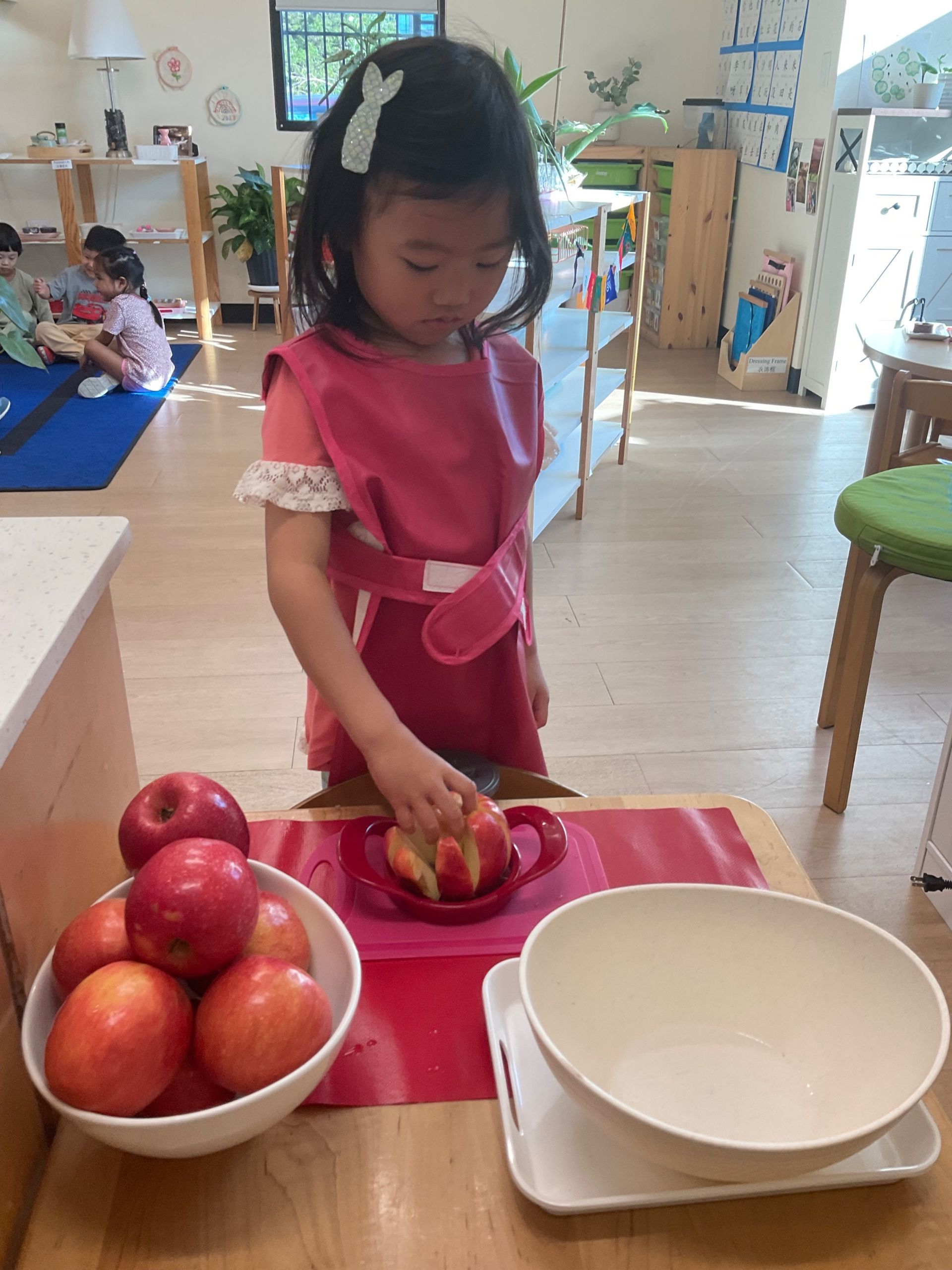 Child in a pink apron arranging fruit on a table in a bright kitchen.